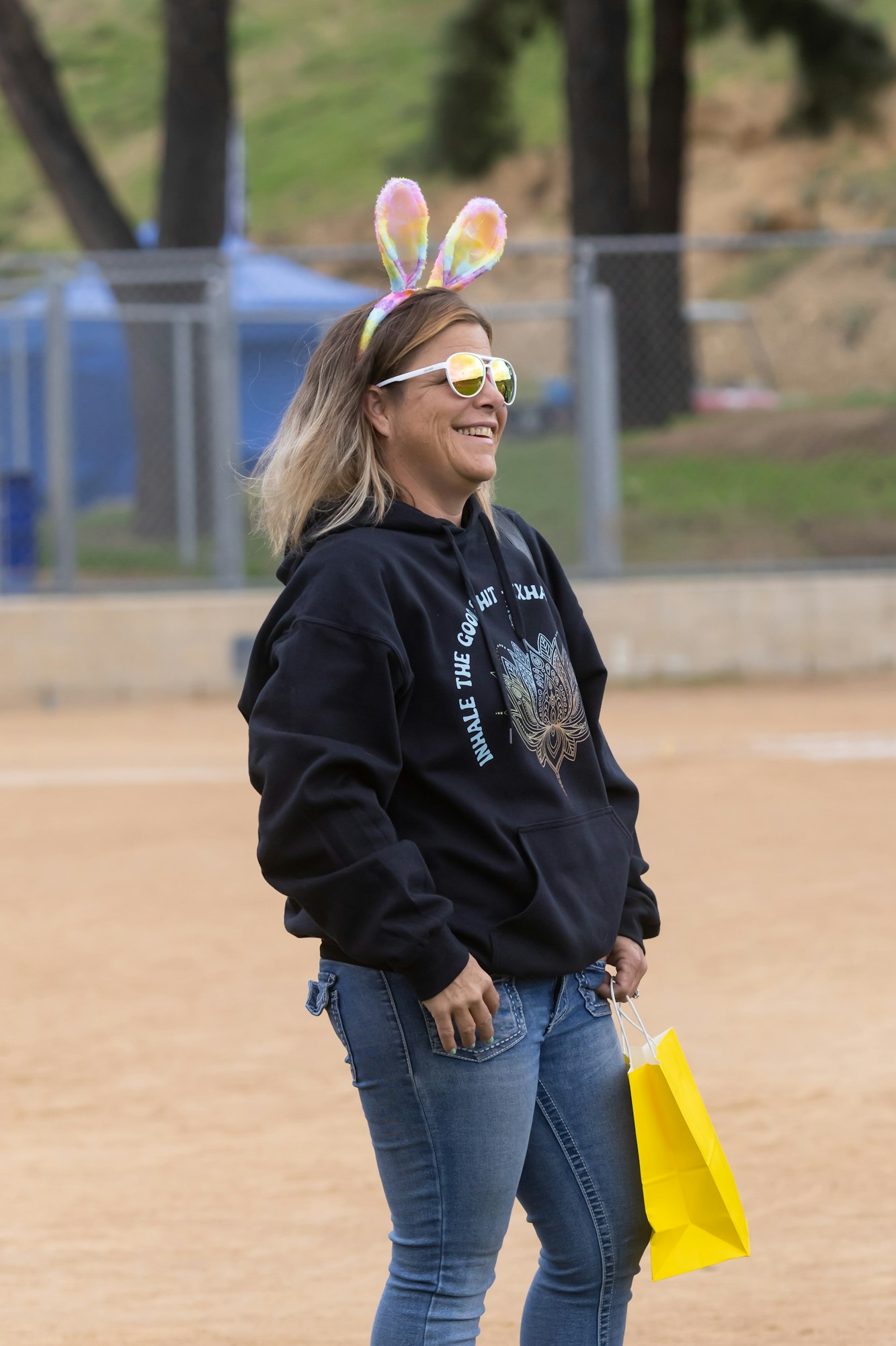 A person with colorful bunny ears and sunglasses, wearing a black hoodie, holds a yellow bag outdoors.