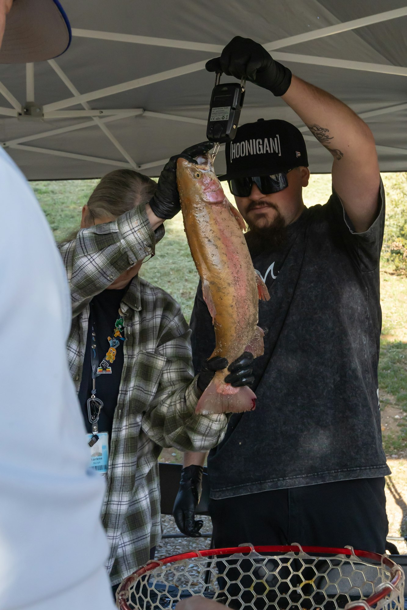 Two people weigh a large fish under a canopy with a digital scale.
