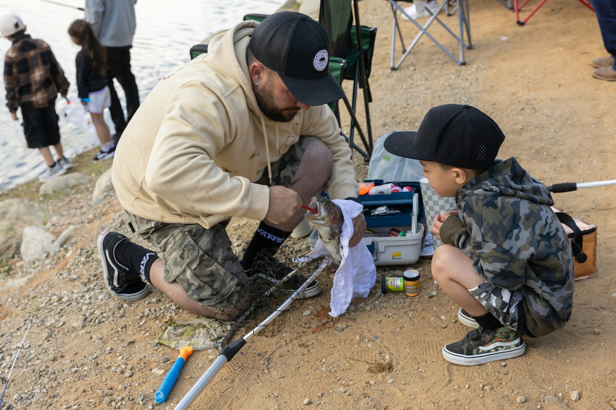 Man shows fish to child at a lakeside, others in background.