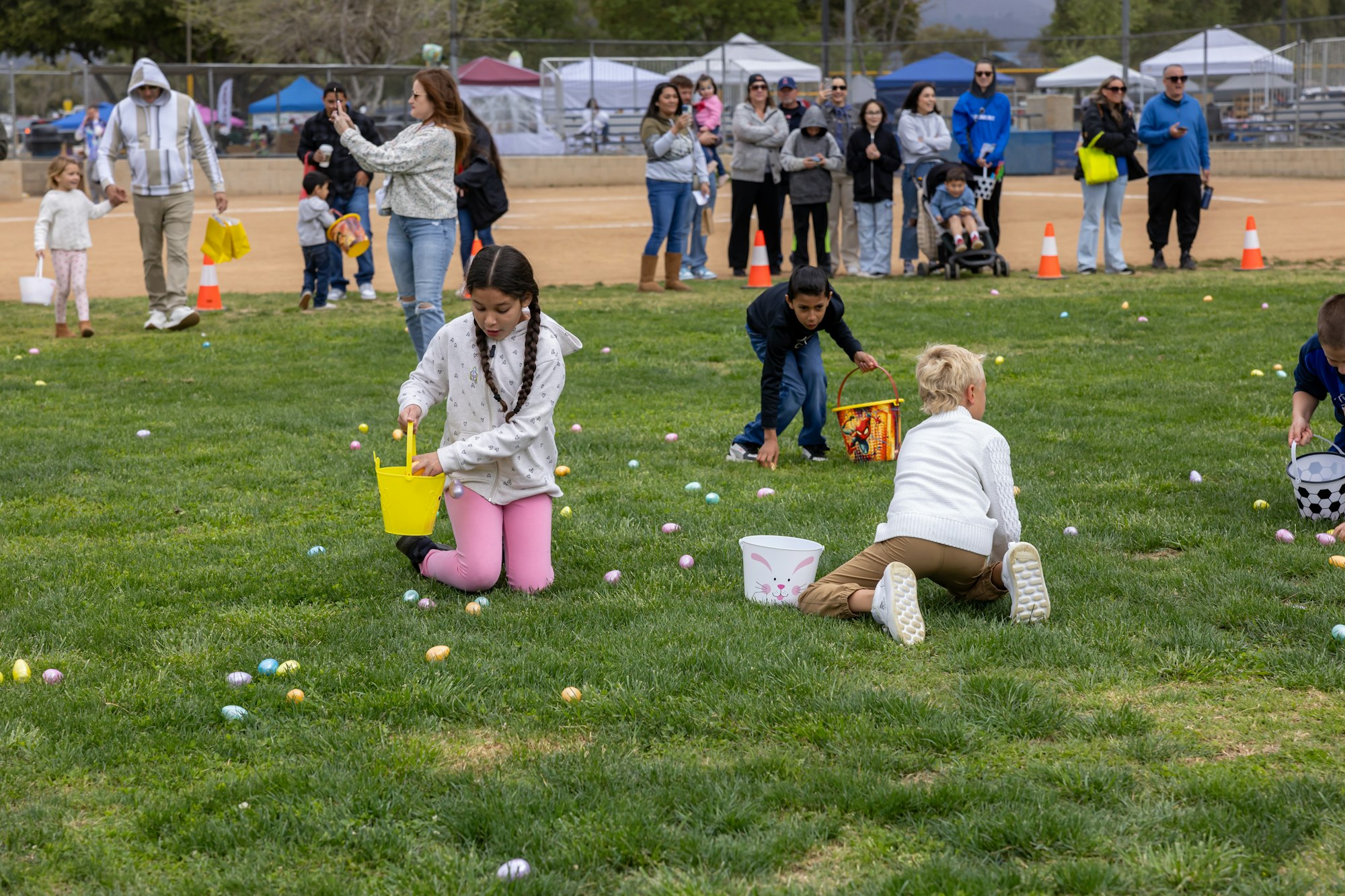 Children collecting eggs in buckets during an Easter egg hunt on a grassy field with adults and other children in the background.