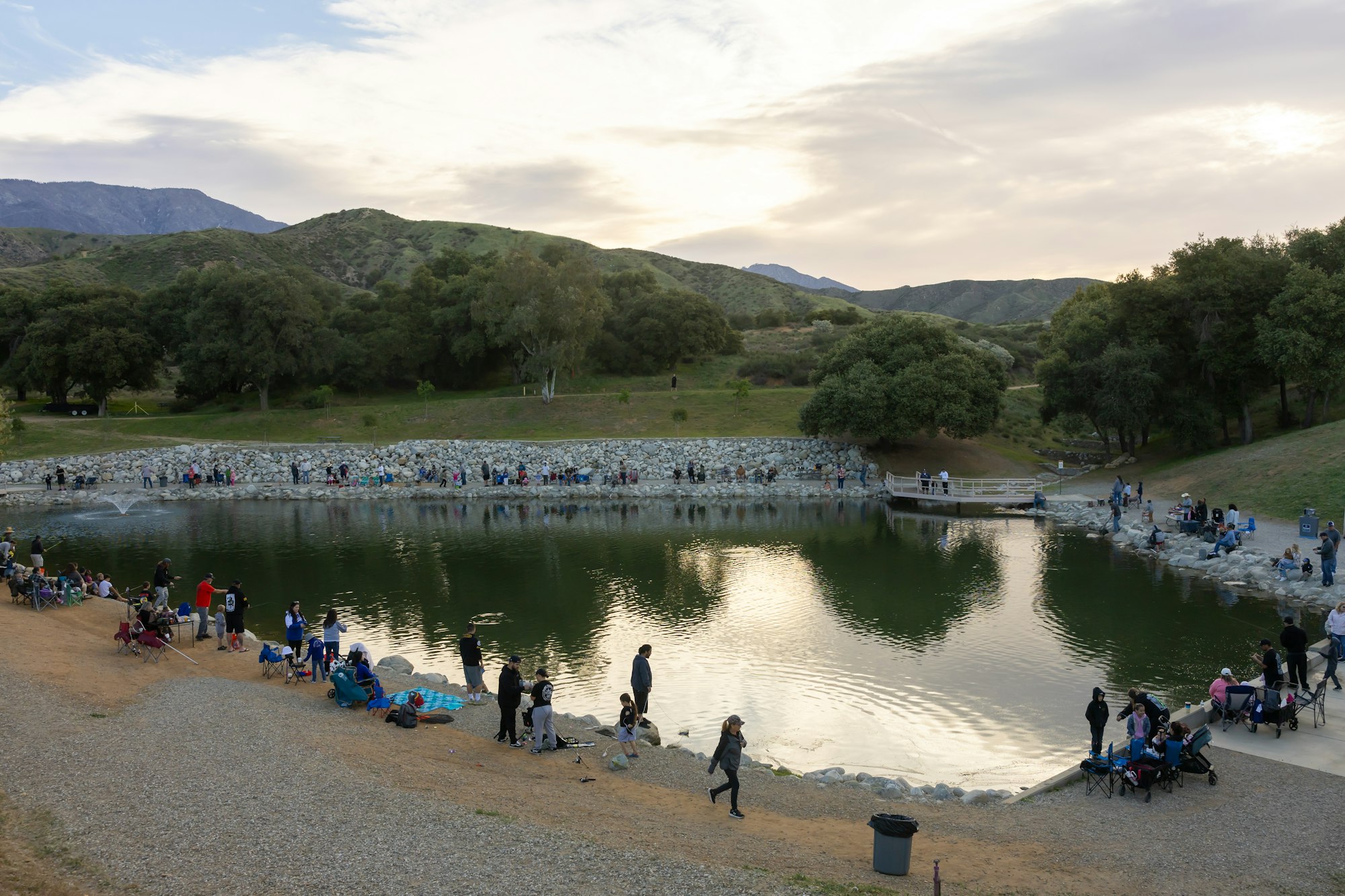 People fishing around a small lake surrounded by hills and trees at dusk.