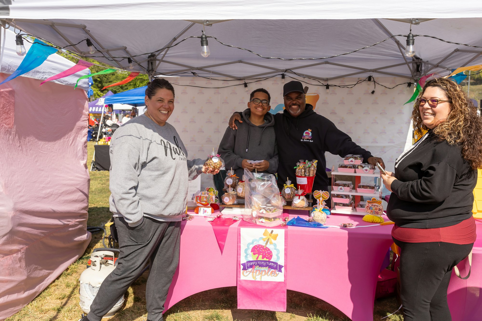 Four people smiling at a booth with treats under a canopy, featuring colorful decorations and a sign for "Yummy Tummy Apples."