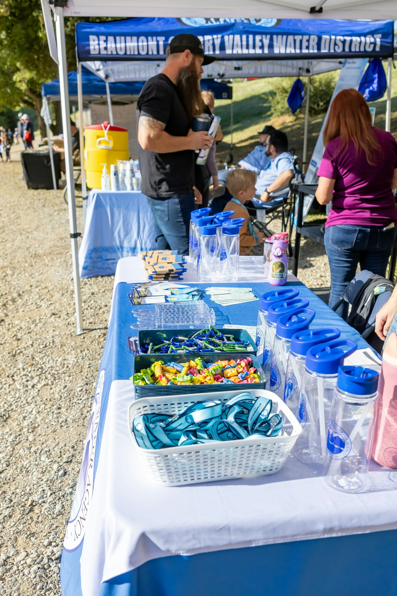 Outdoor event booth with water bottles, lanyards, and candies on a table, people gathered around.