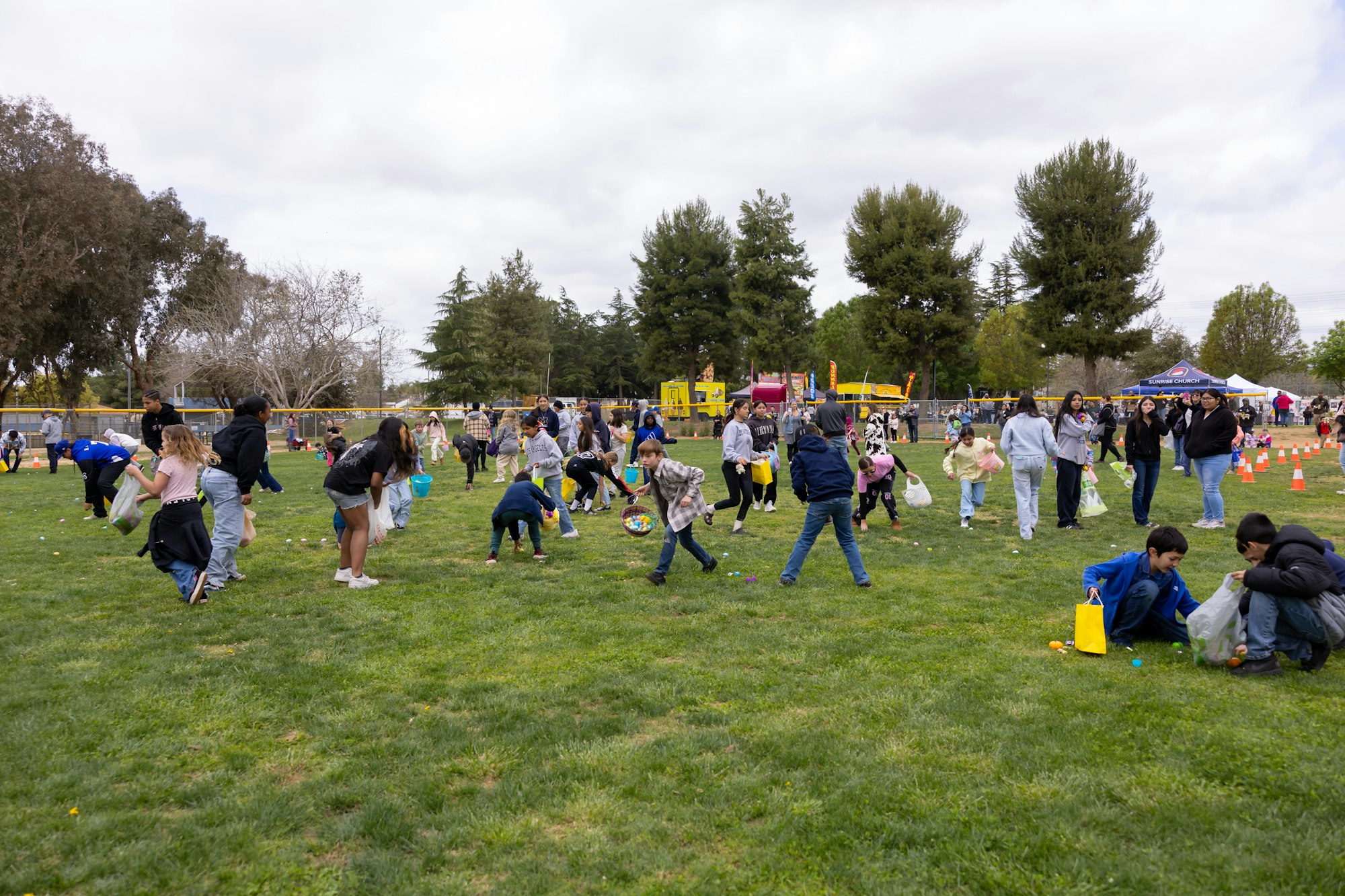 People participating in an outdoor event, possibly an egg hunt, with children collecting items in bags on a grassy field.