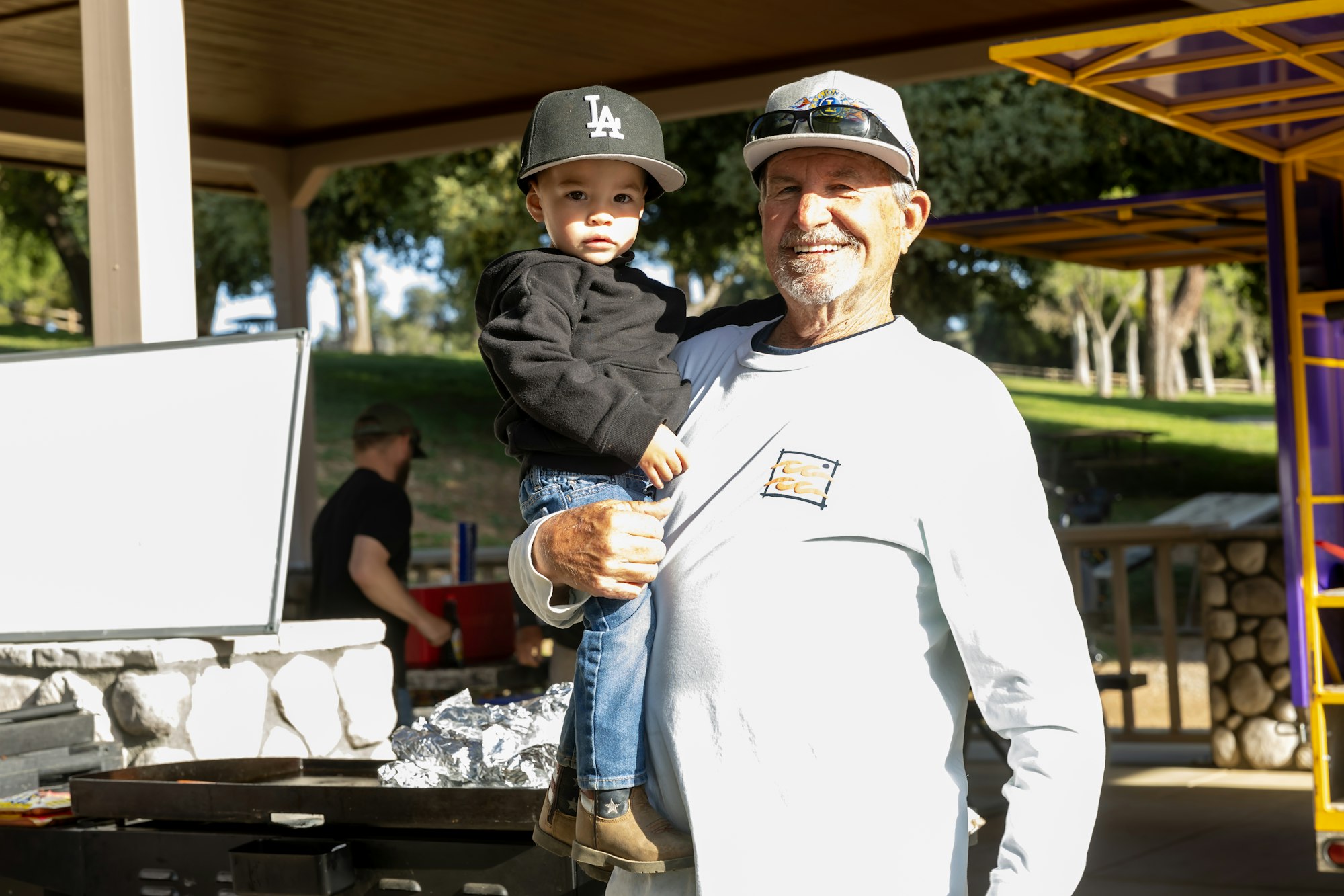 A smiling man holds a child under a pavilion, surrounded by picnic tables and trees.