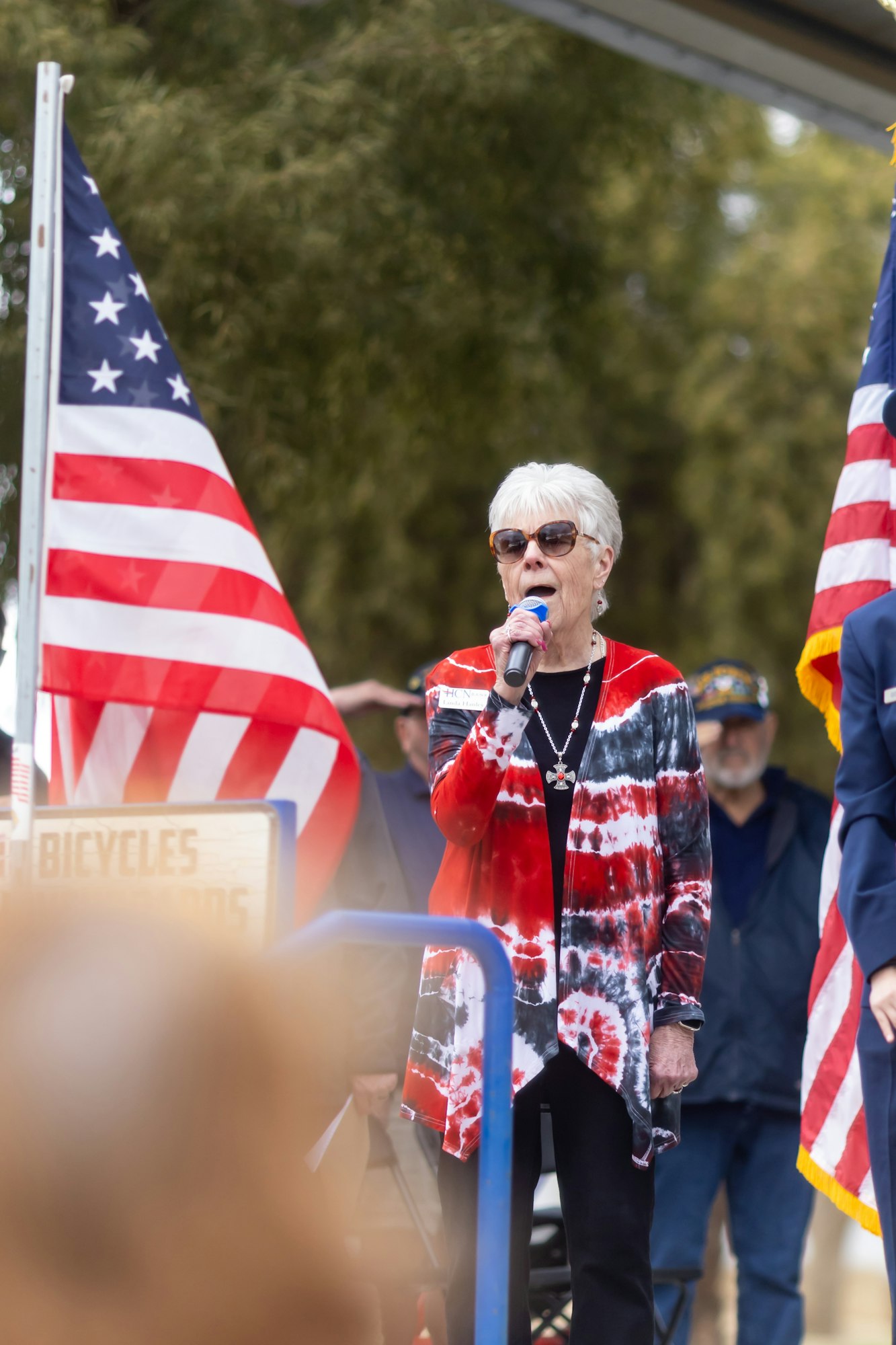 A person with sunglasses holds a microphone, standing next to American flags, possibly at a public event or ceremony.
