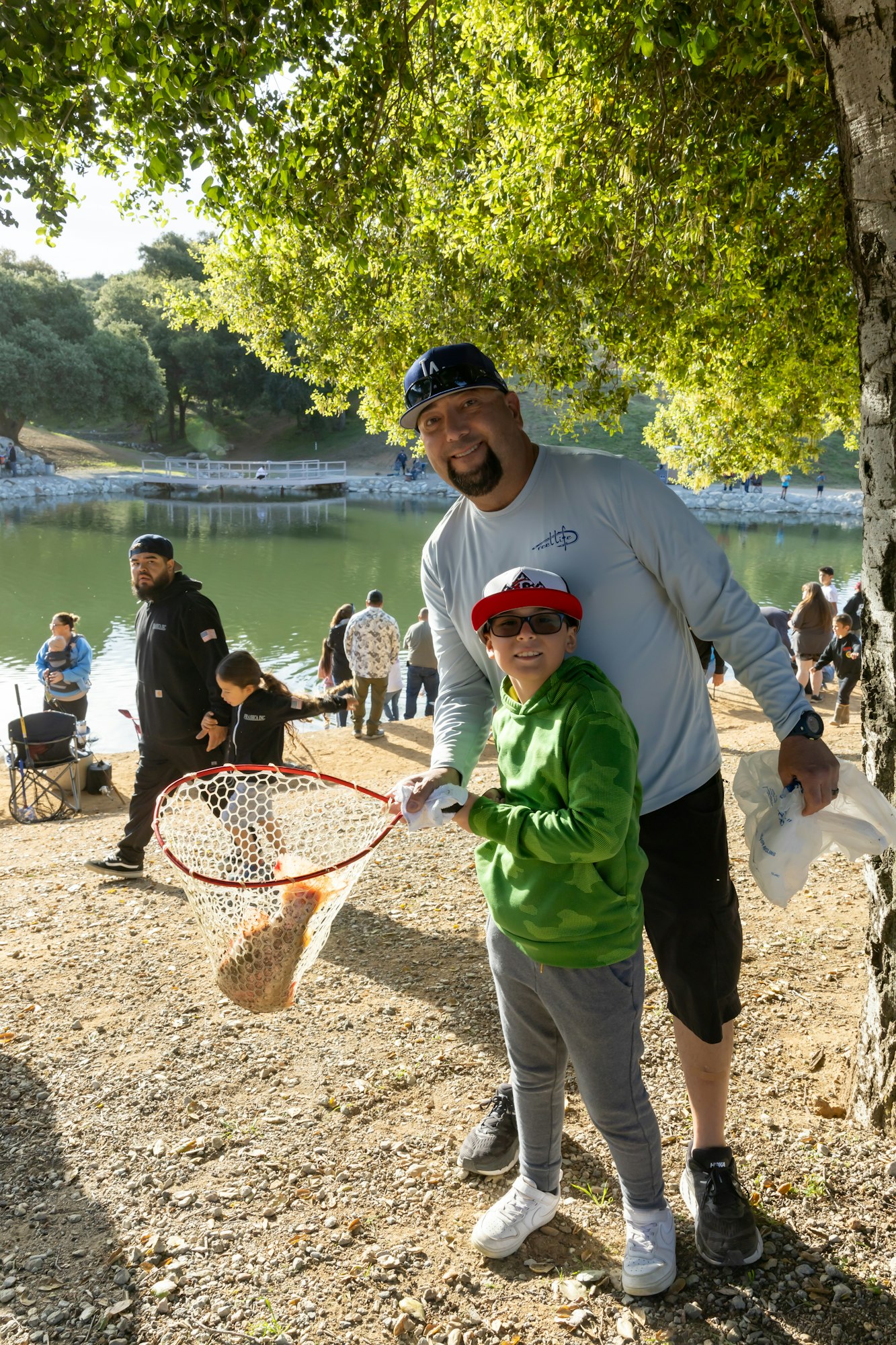 A man and child smile while holding a fishing net with a fish. They're near a lake with others in the background.