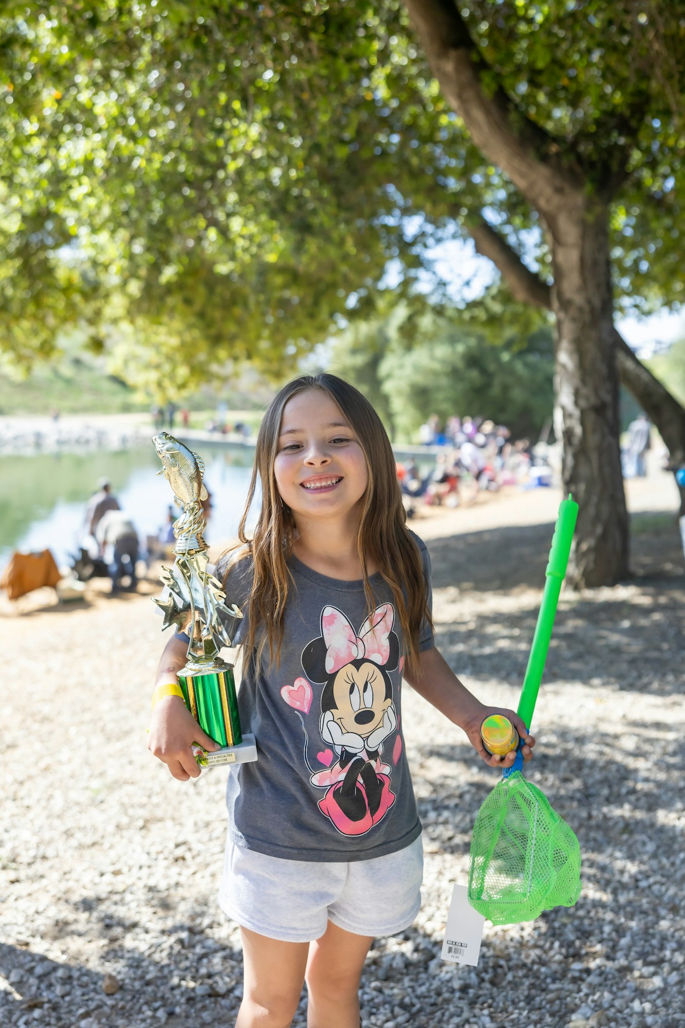 A smiling girl holding a fishing trophy, green net, and bubble wand, wearing a Minnie Mouse shirt, outdoors by a pond.