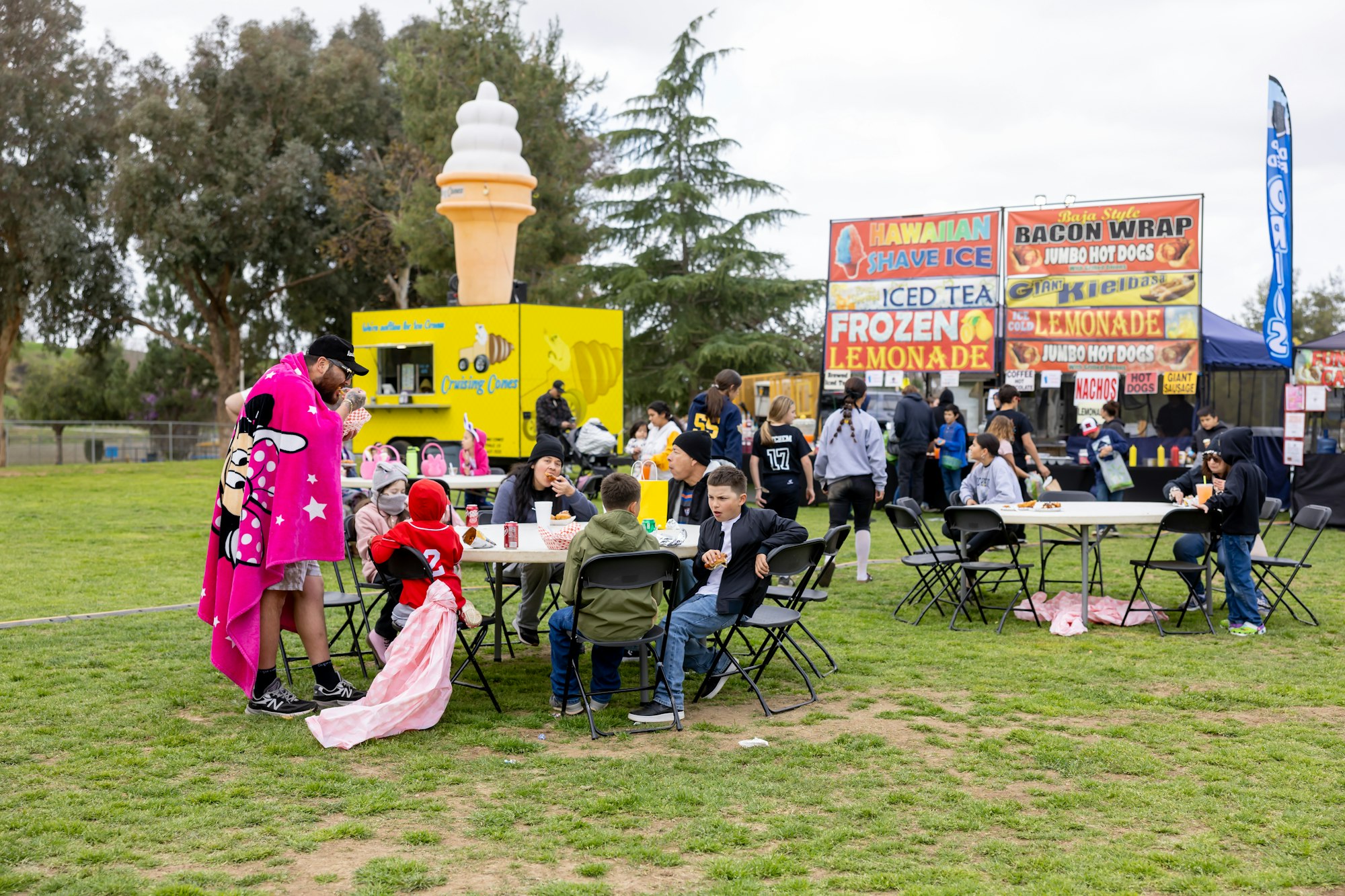 People at an outdoor event with food stalls, tables, and a large ice cream cone sign.
