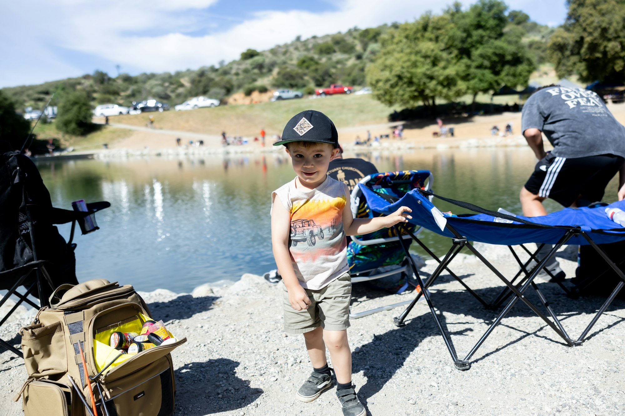A child in a cap stands near a lake with folding chairs, a backpack, and people in the background.