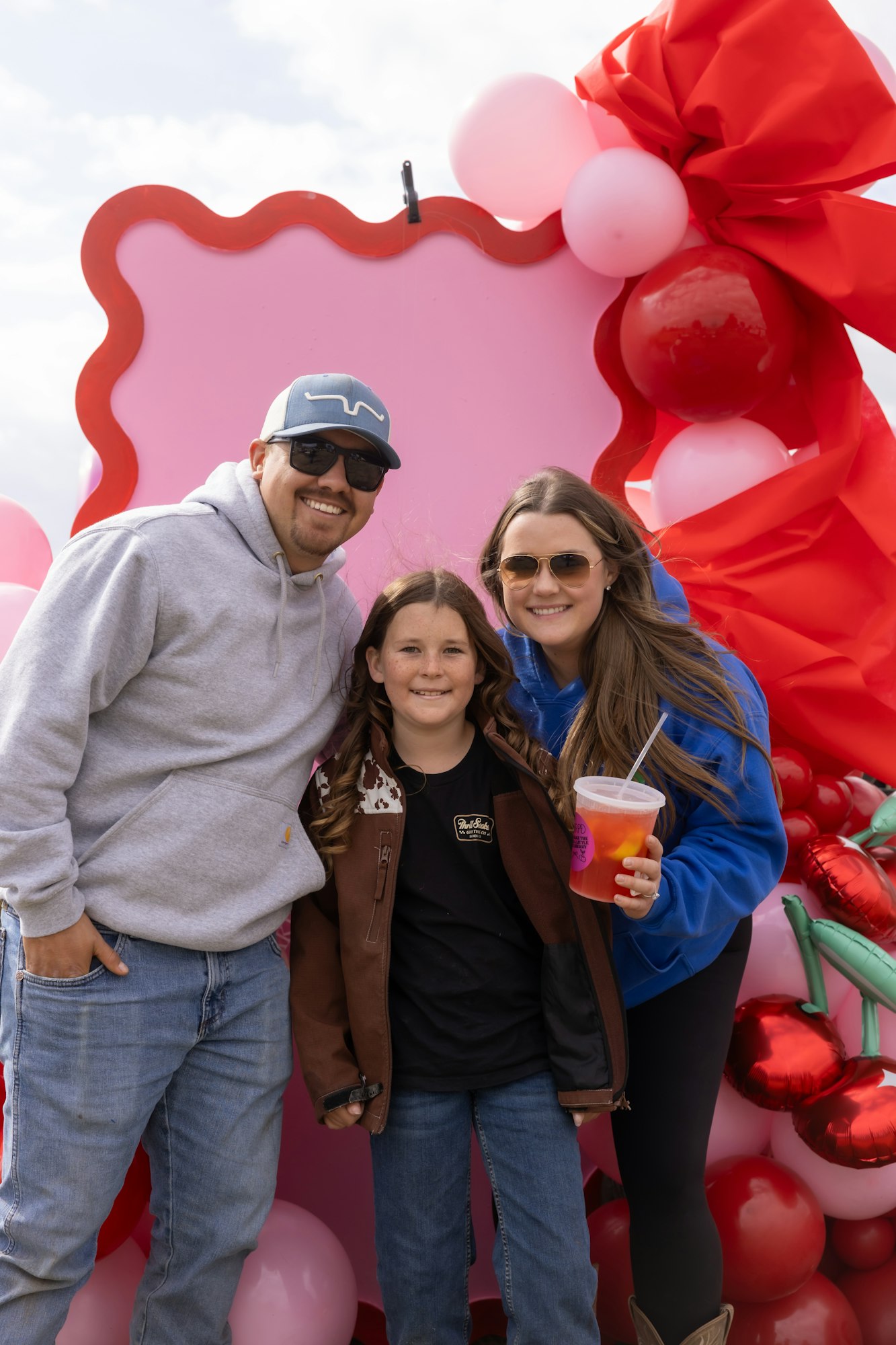 Three people smiling in front of a pink backdrop with balloons and a large red bow.