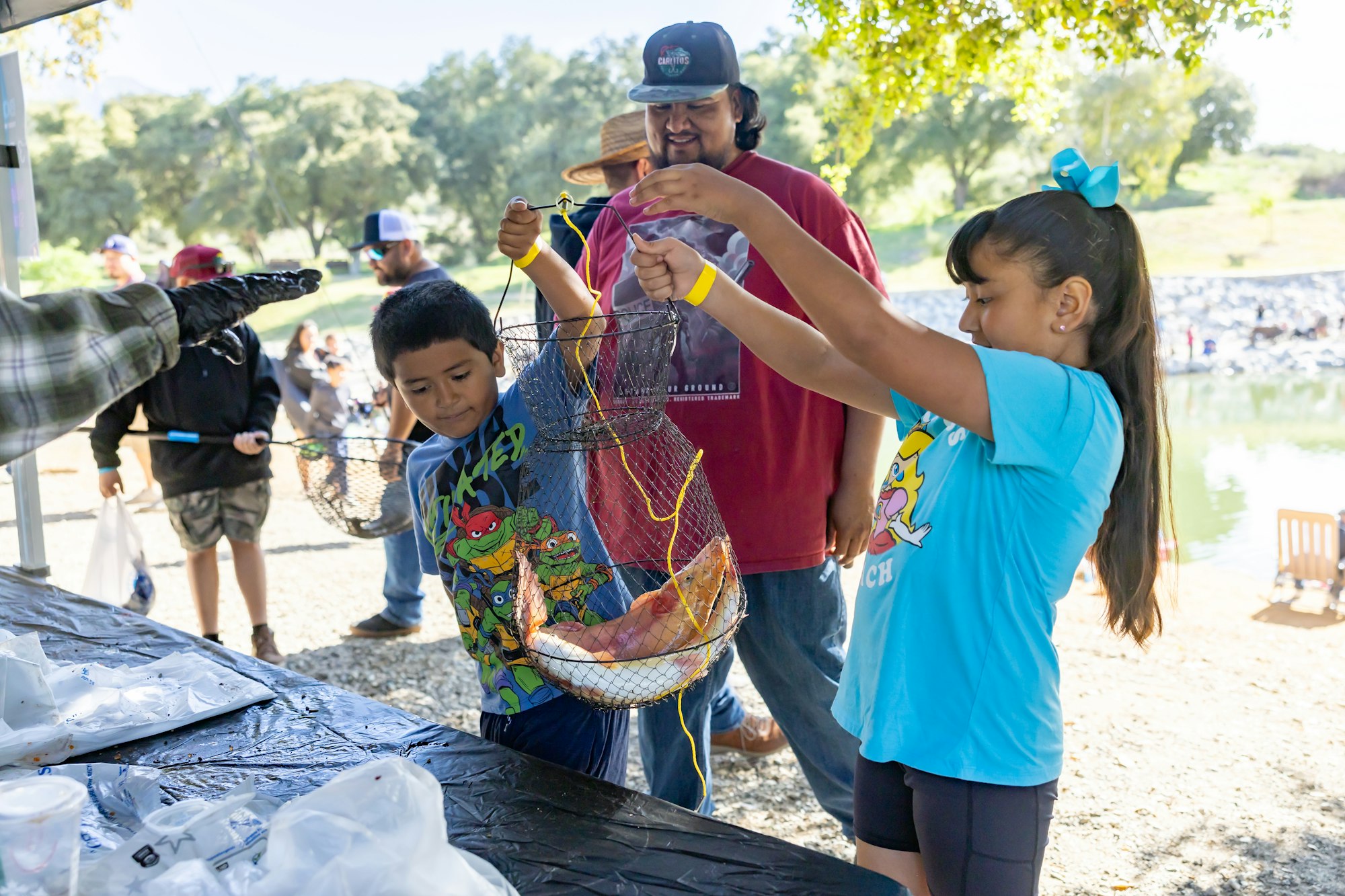 Kids show fish in a net at an outdoor event, with adults nearby and a lake in the background.