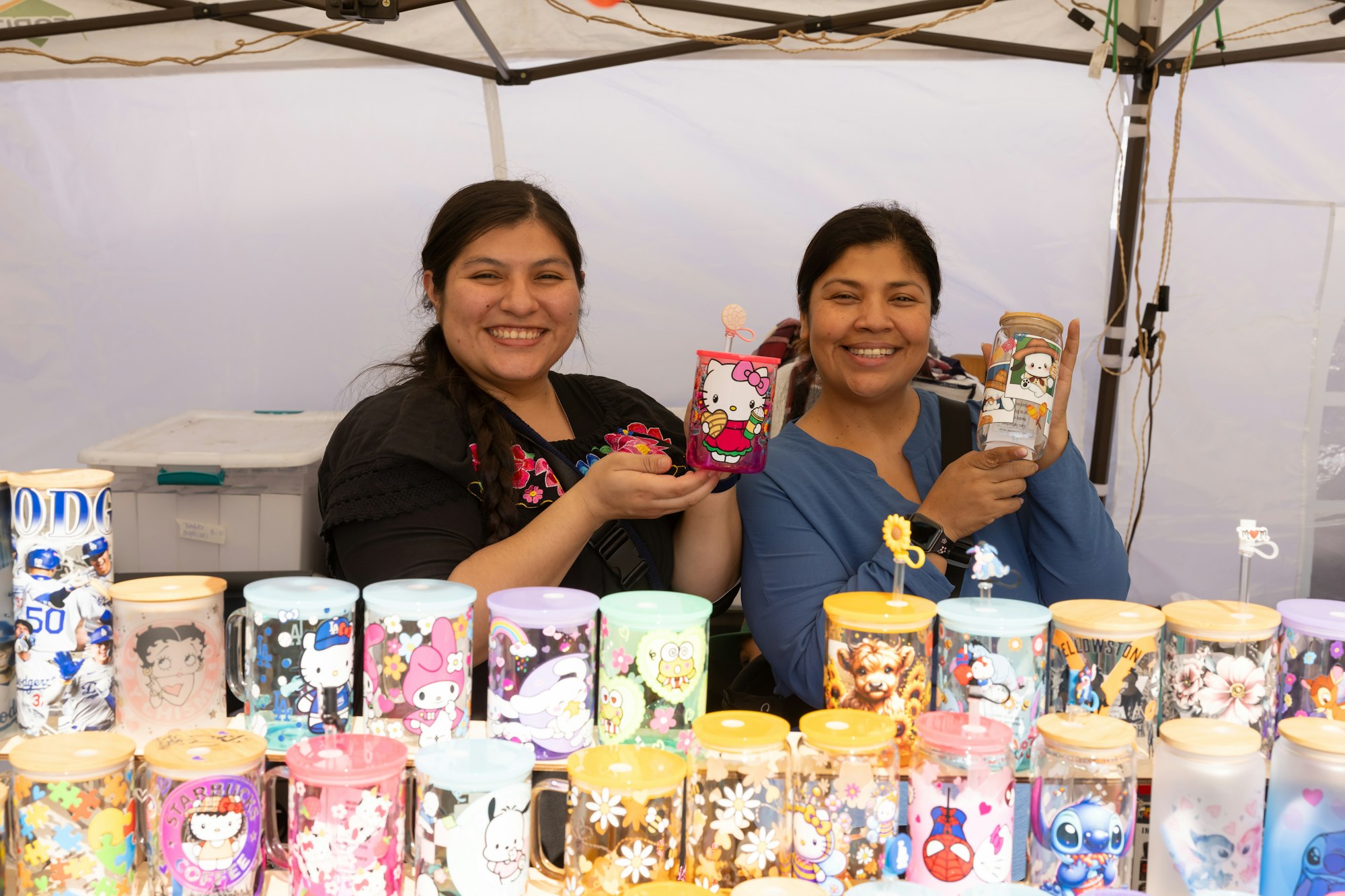 Two people smiling, displaying colorful cups with cartoon designs at a market stall.