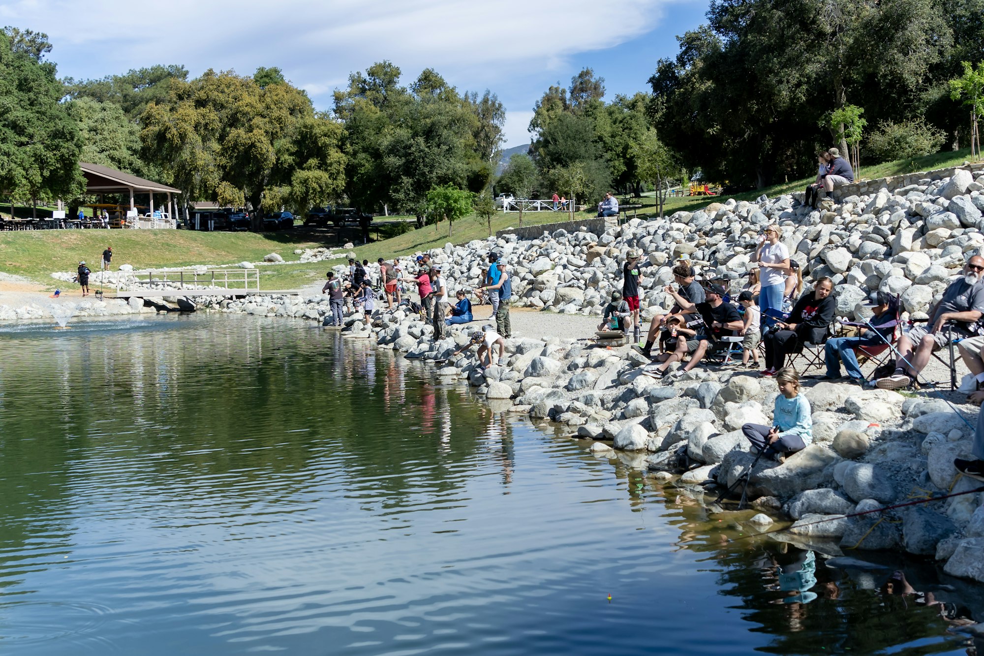 People fishing along a rocky shoreline with trees and a pavilion in the background.