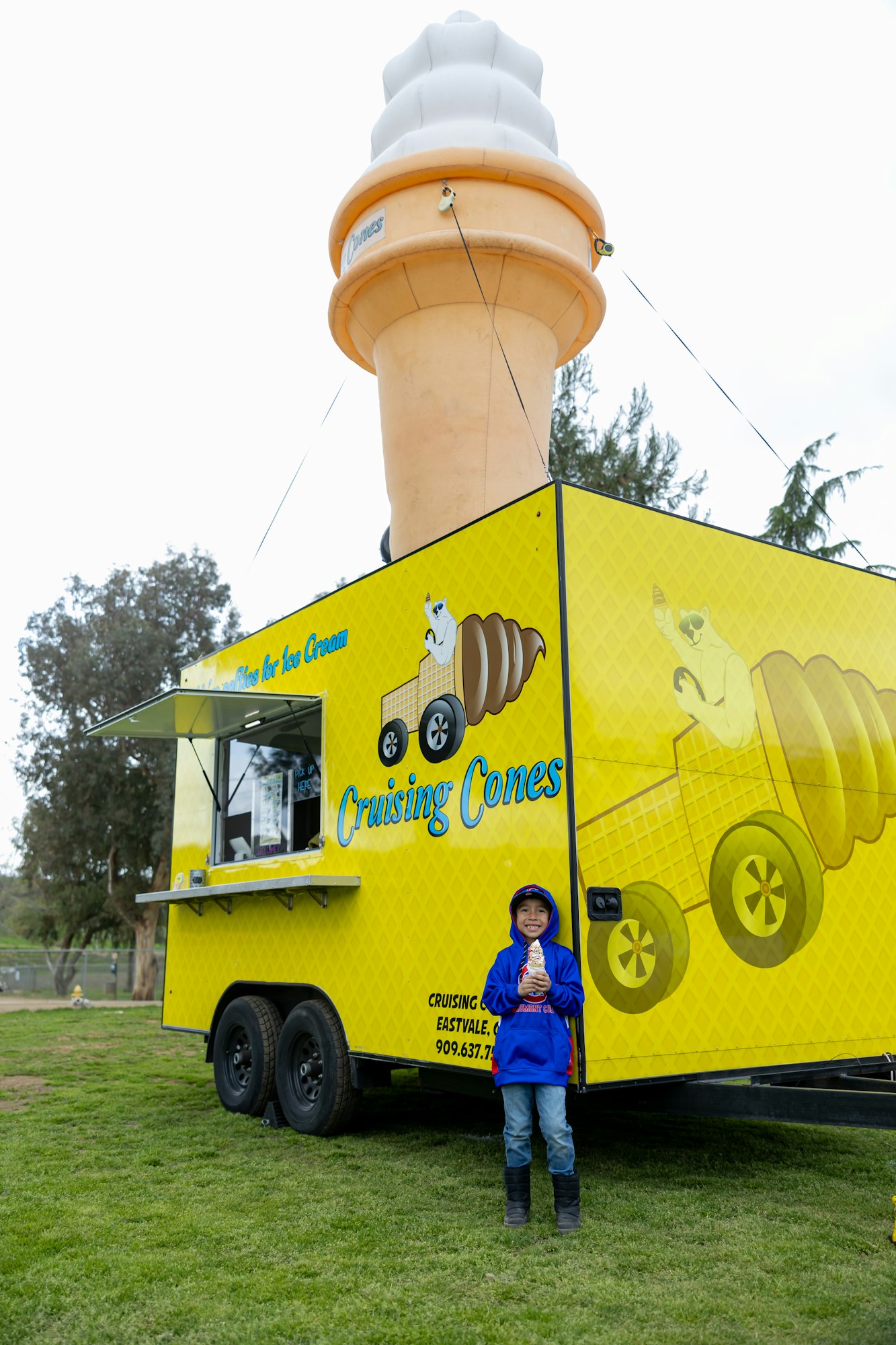 A child holding ice cream stands beside a yellow "Cruising Cones" truck with a large ice cream cone model on top.