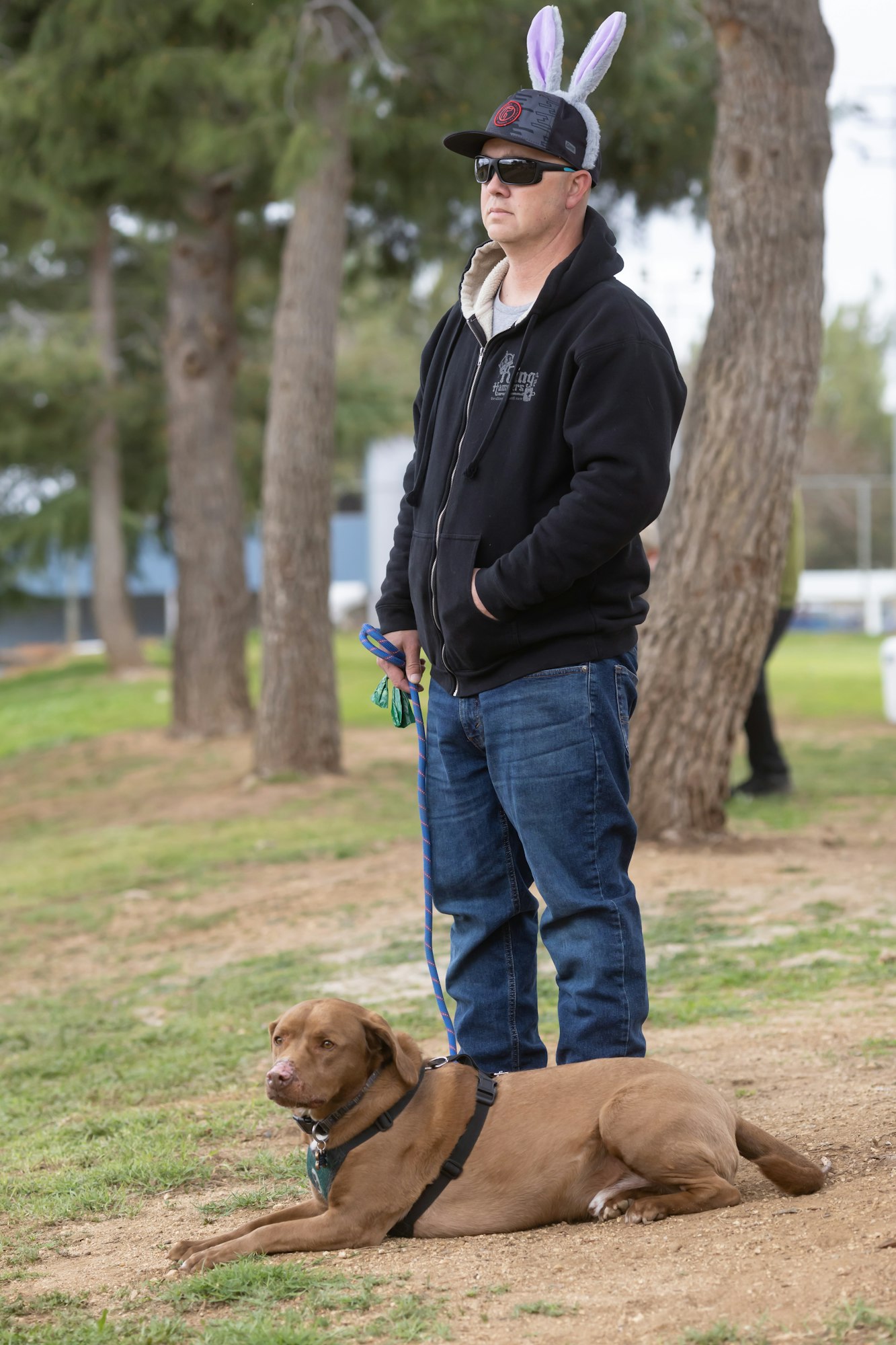 A person wearing bunny ears stands with a dog on a leash in a park setting.