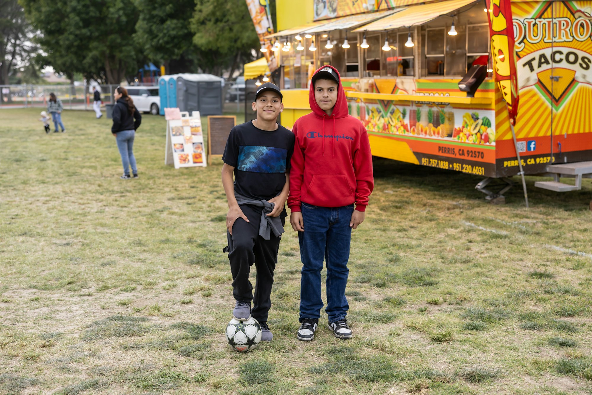 Two boys stand on grass near a taco truck; one has a soccer ball.