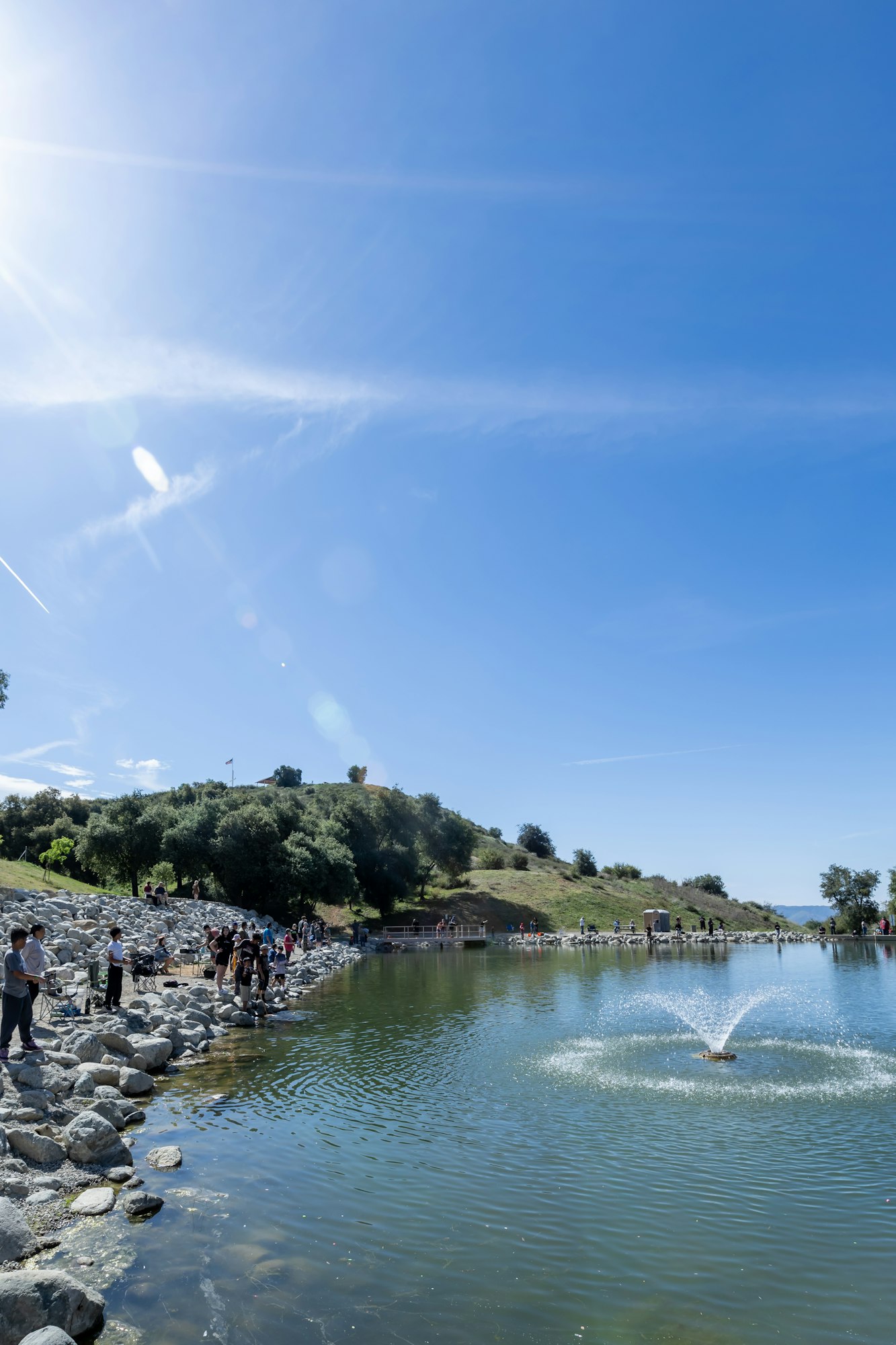 A scenic view of a lake with a fountain, people on the rocky shore, a green hillside, and a bright blue sky above.