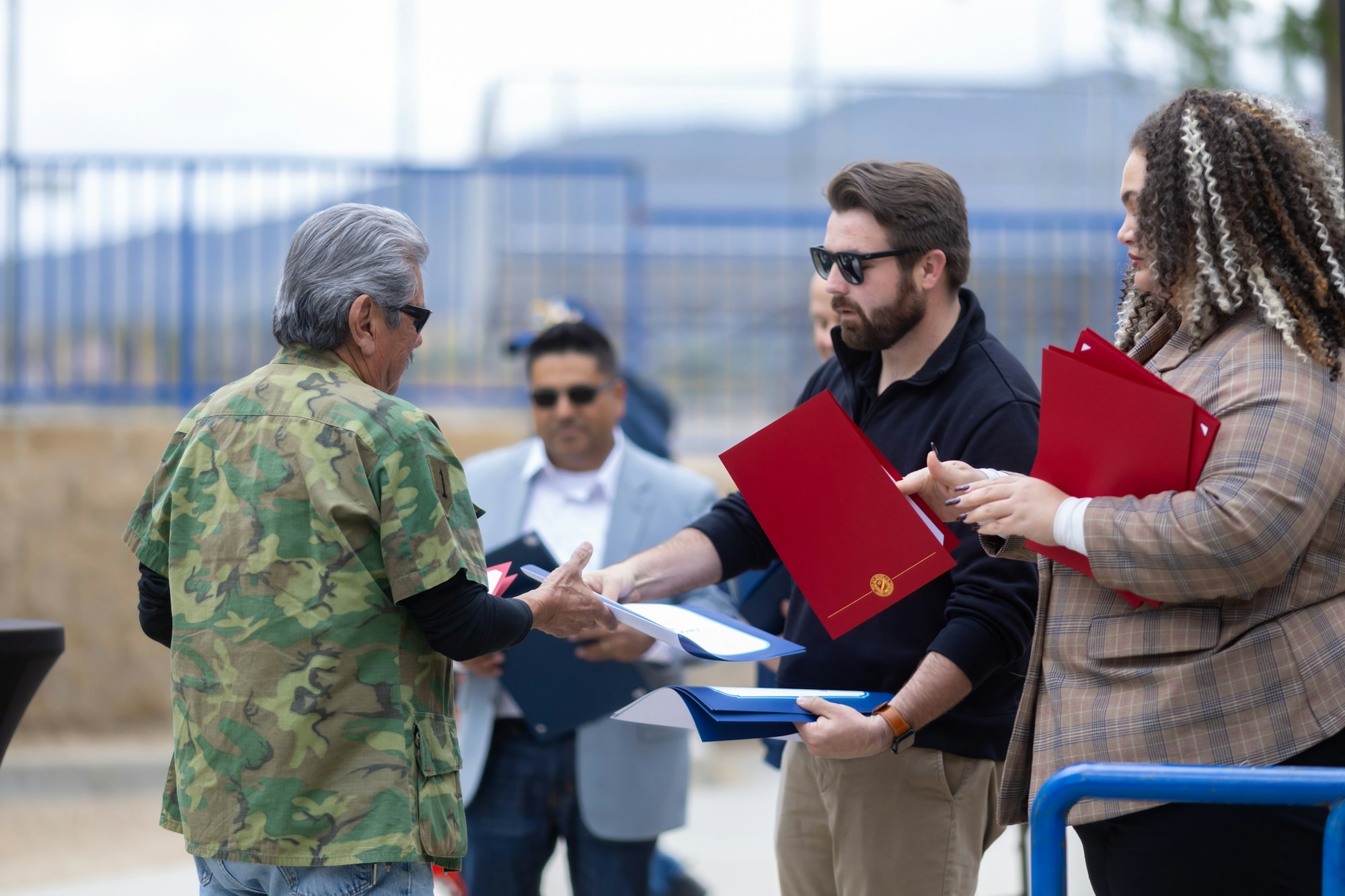 People exchanging documents outdoors, one in camo, the others holding red folders.
