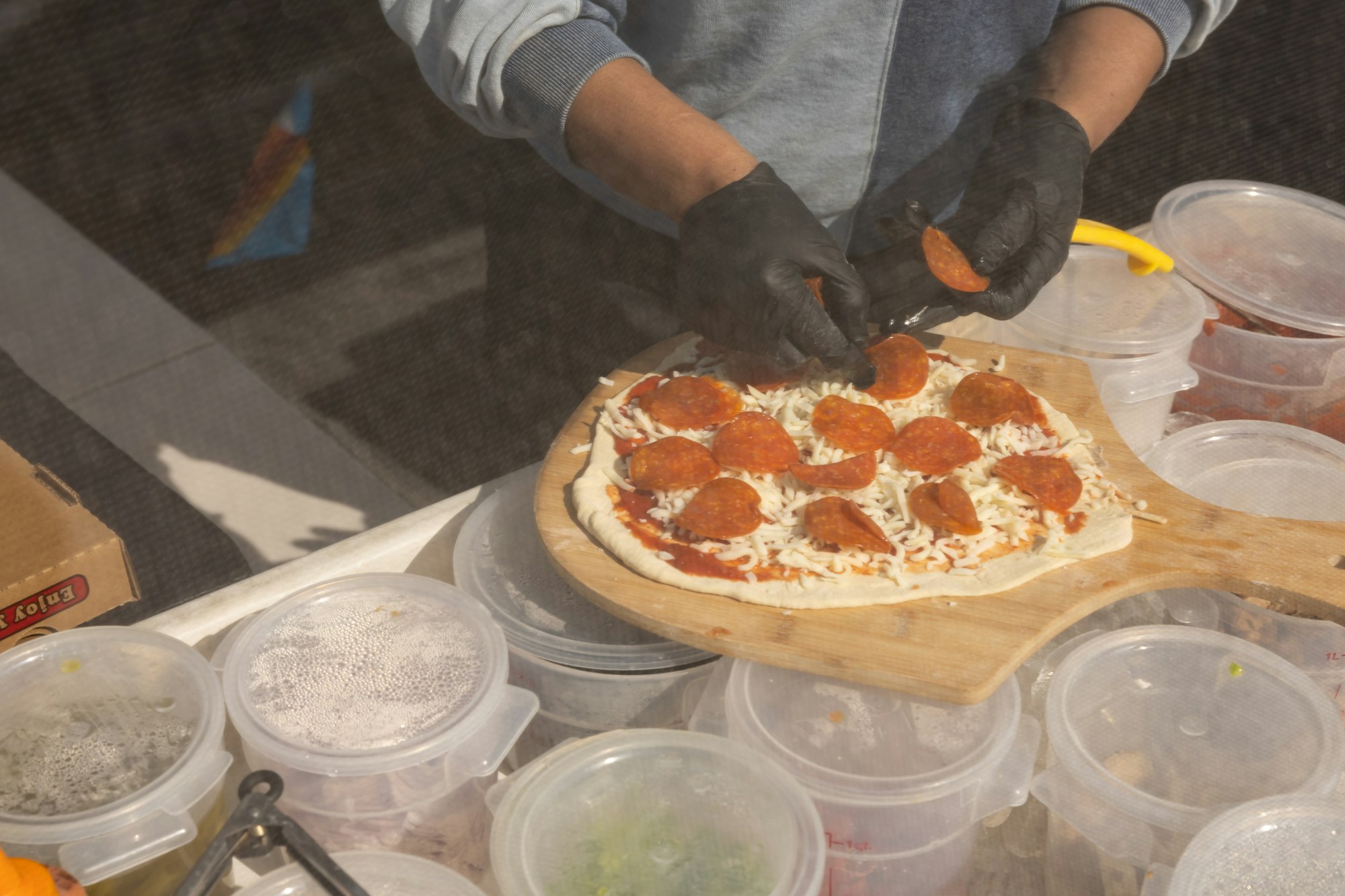 Person making a pepperoni pizza, adding toppings on dough with cheese and sauce, surrounded by containers of ingredients.