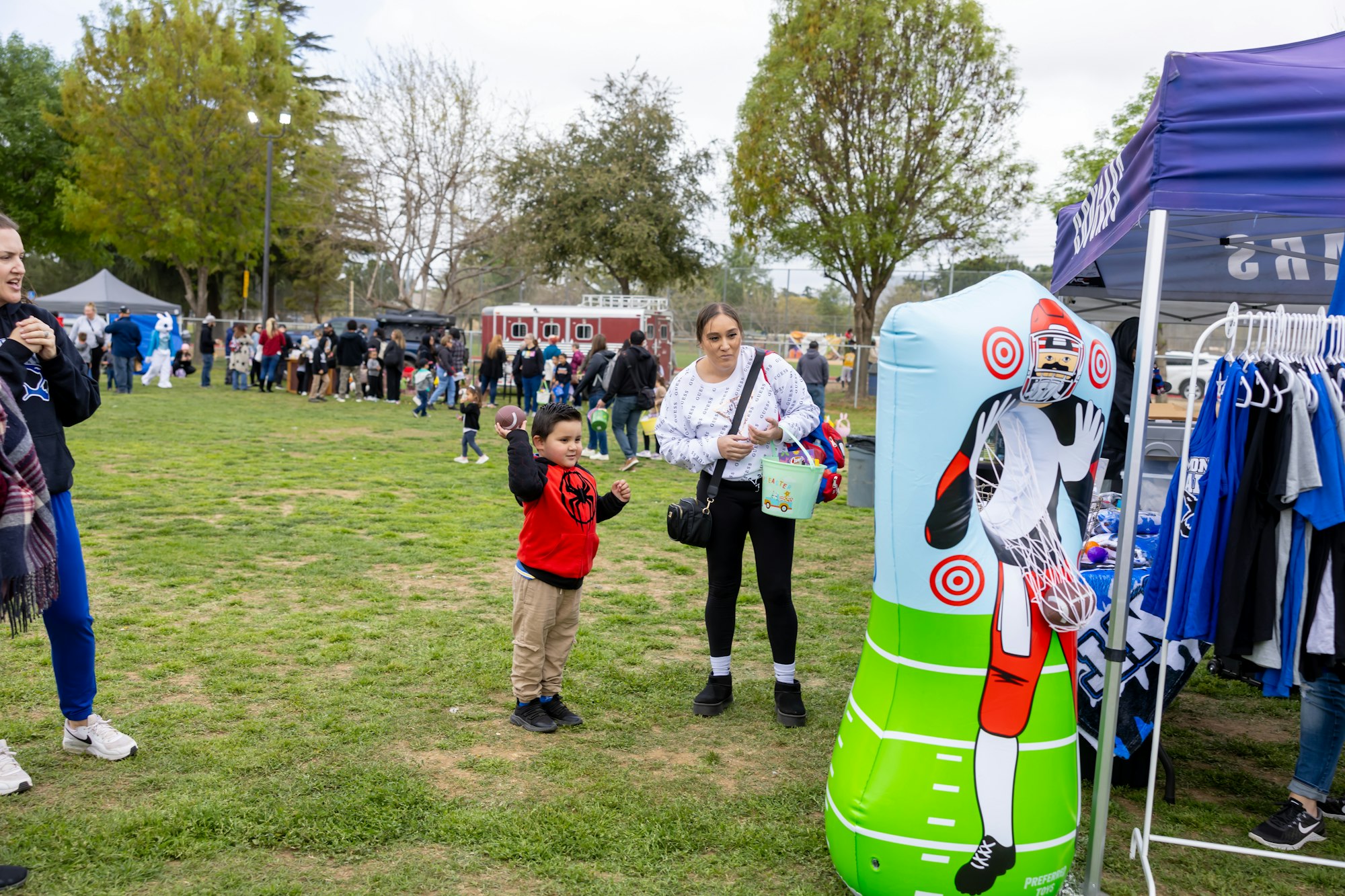A child aims a football at an inflatable target game at an outdoor event, with people and booths in the background.