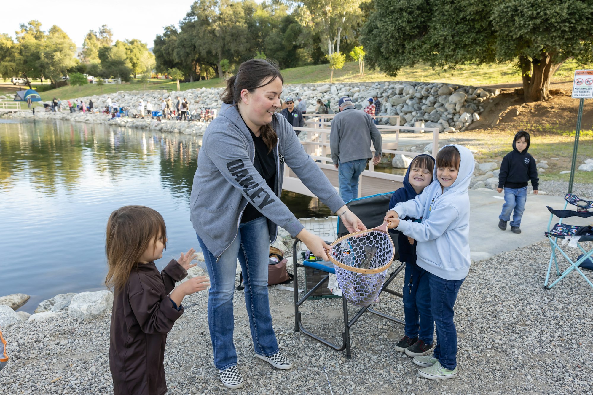 A woman and two children hold a fishing net with a fish by a lake; others fish in the background.