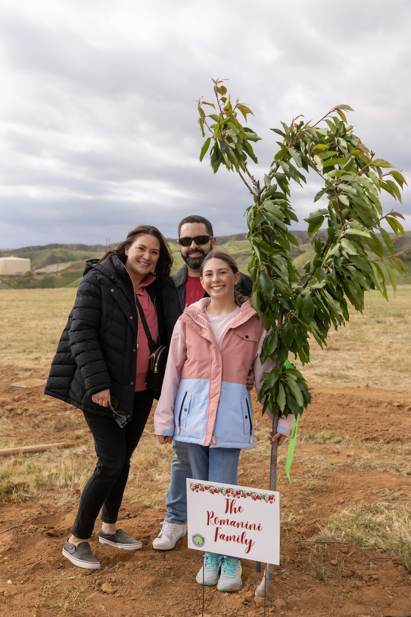 A family of three posing with a small tree and a sign reading "The Romanini Family" in a field.