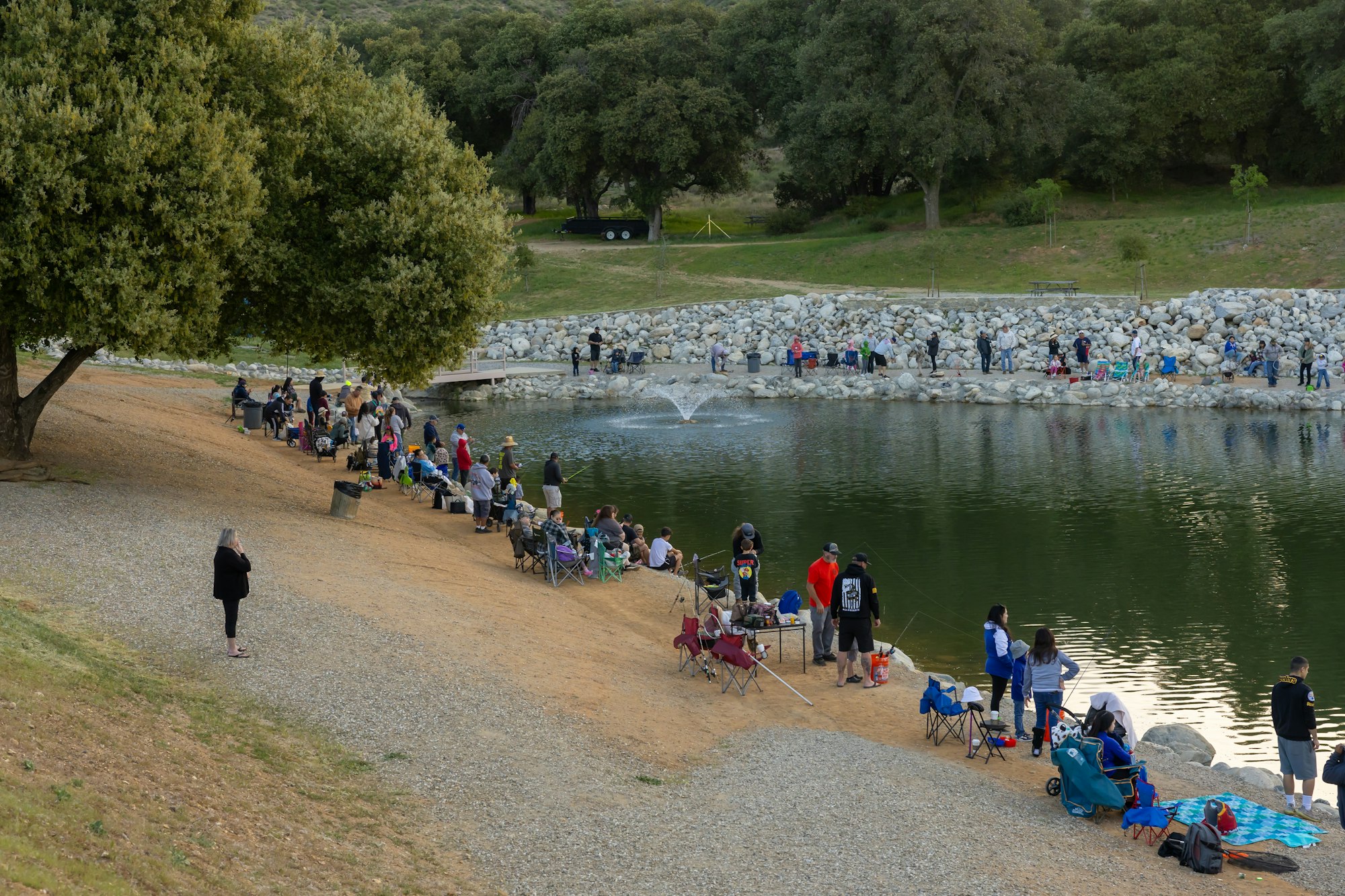 People are fishing and relaxing by a large pond, surrounded by trees and rocks, with a fountain in the center.