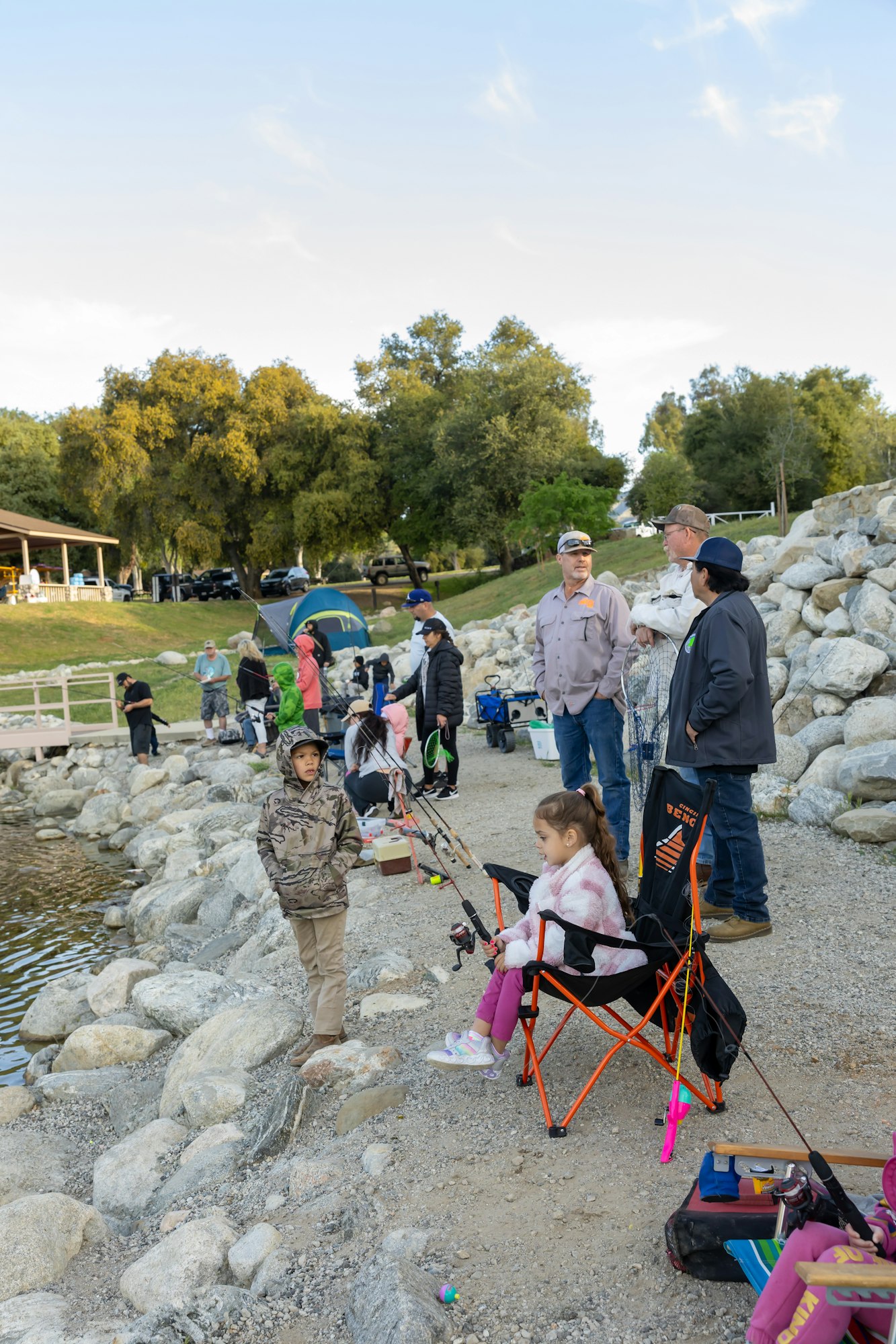 People fishing by a lakeside, children and adults gathered with gear, trees in background.