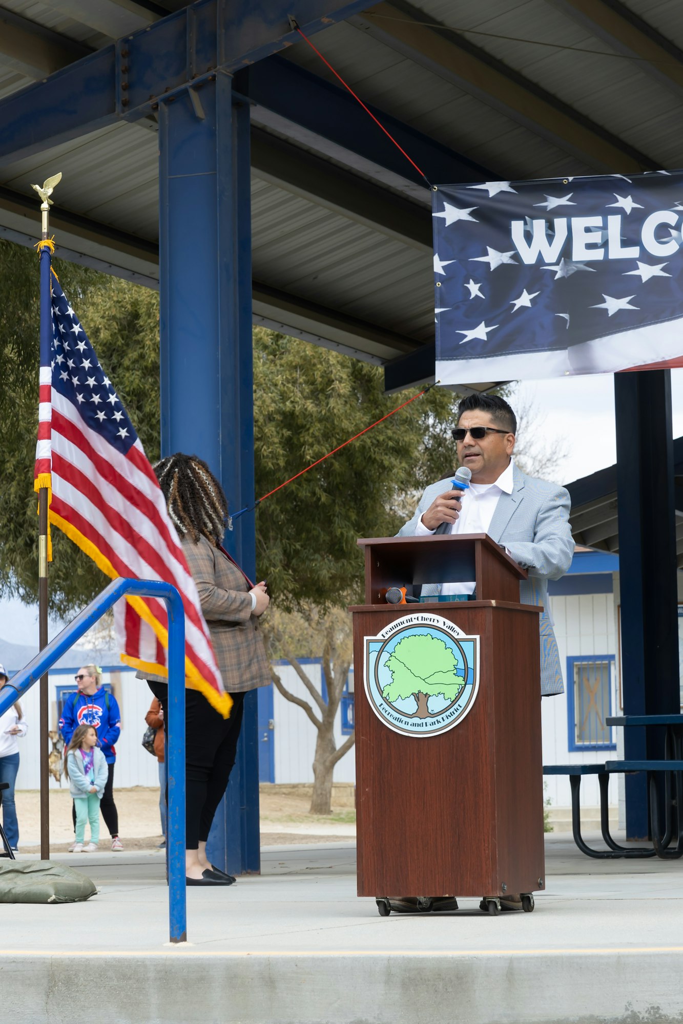 A person speaking at a podium with a microphone, an American flag, and a "WELCOME" banner overhead.