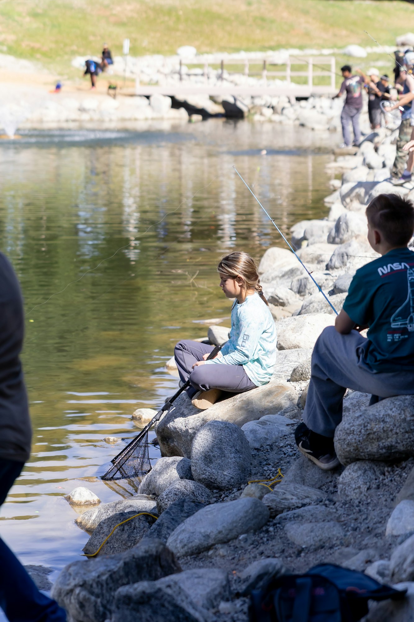 Children fishing by a rocky lakeside, one holding a net, surrounded by other people with rods.