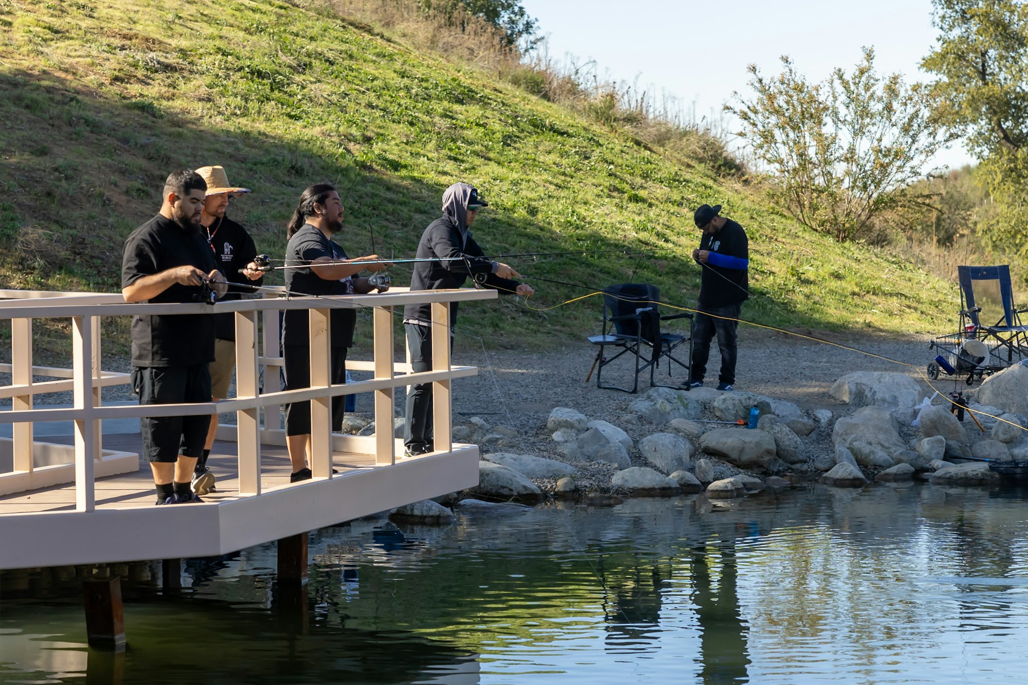 Group of people fishing from a dock and shore by a grassy hill.