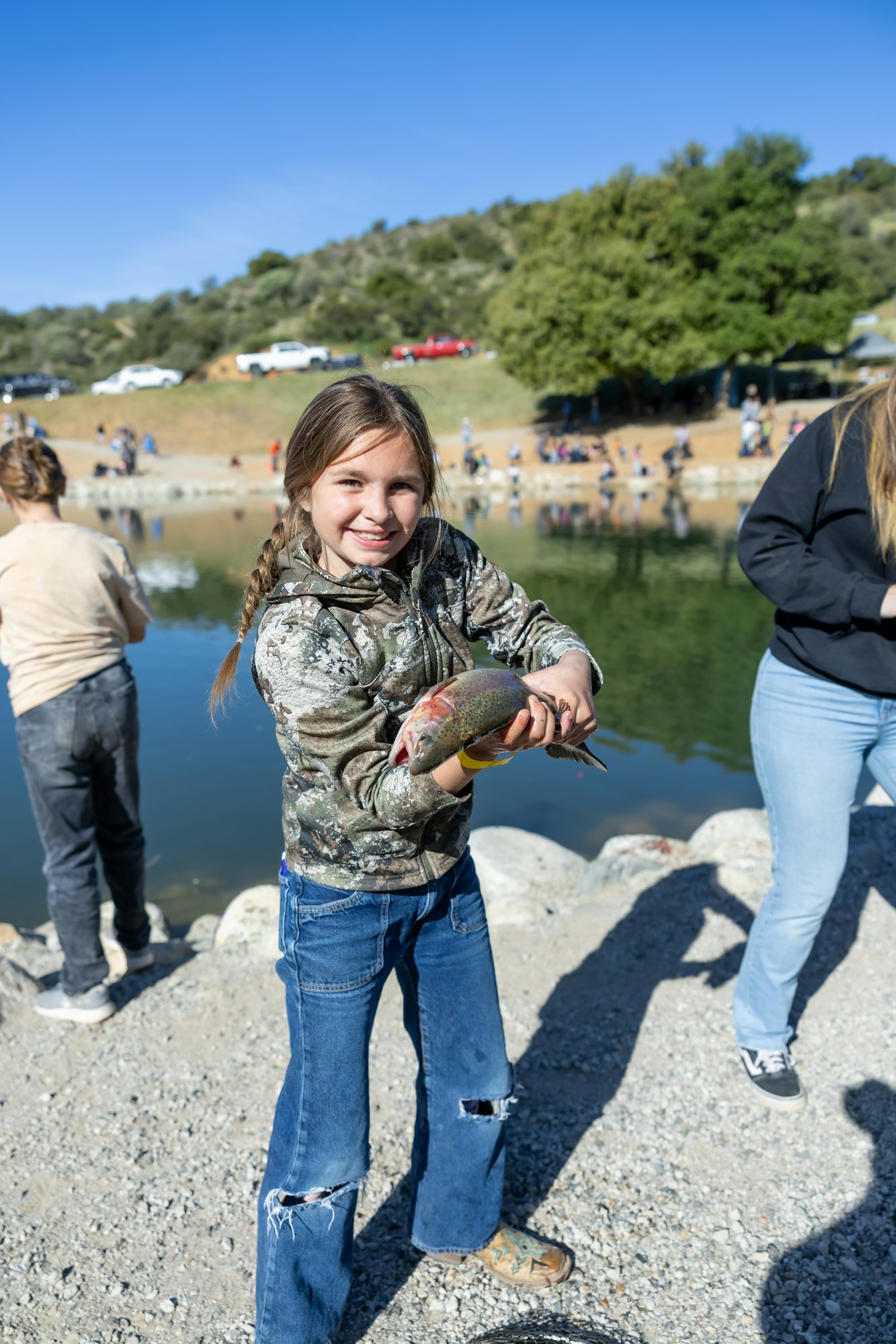 A girl holding a fish by a pond, smiling, with people and cars in the background.