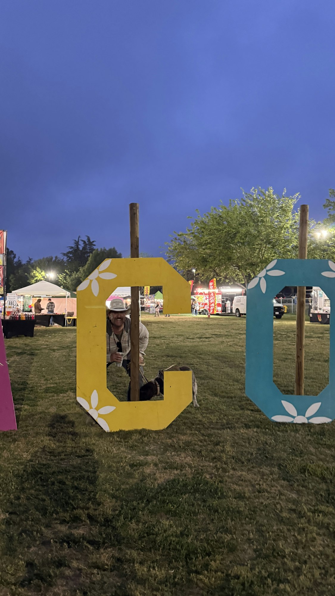 A person peeks through a large "C" sign with floral decor in a park setting, holding a drink, near food stalls at dusk.