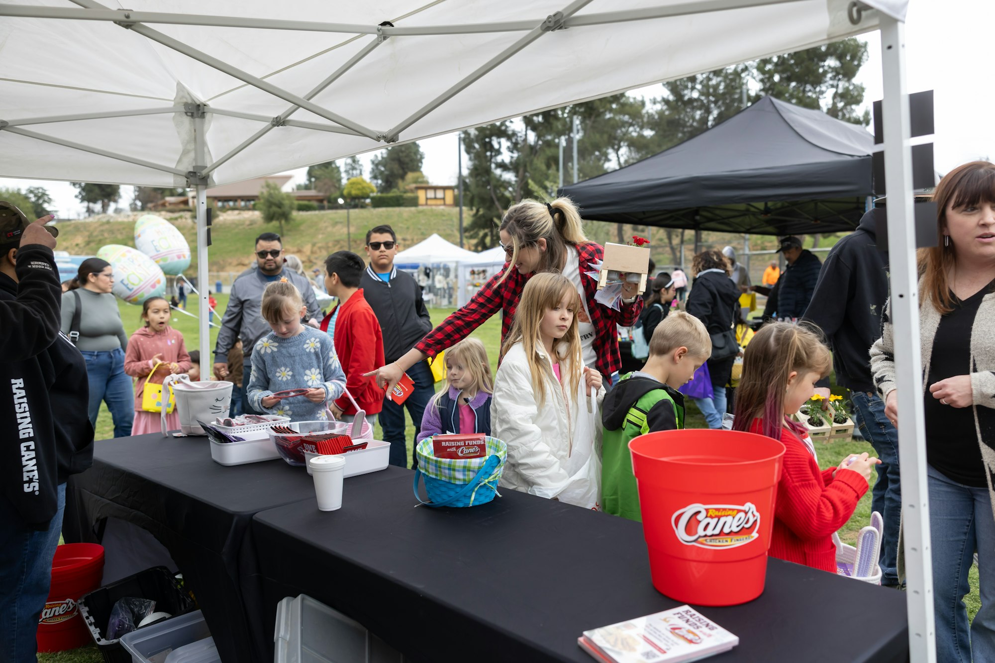 People gathering under tents at an outdoor event, with children holding baskets near a table with branded buckets.