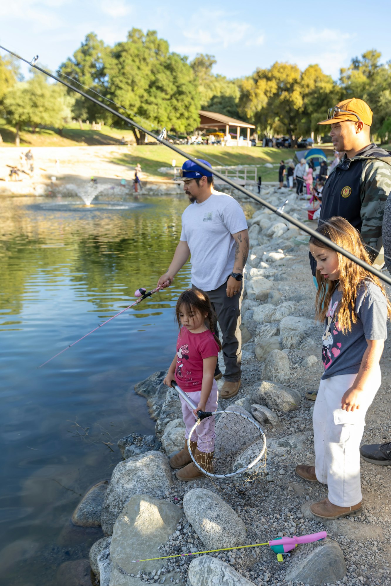 People fishing by a pond, including adults and children with fishing rods and a net.