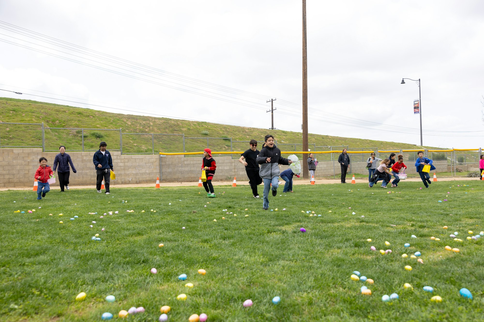 Children running in a field, collecting colorful eggs, with adults watching nearby.