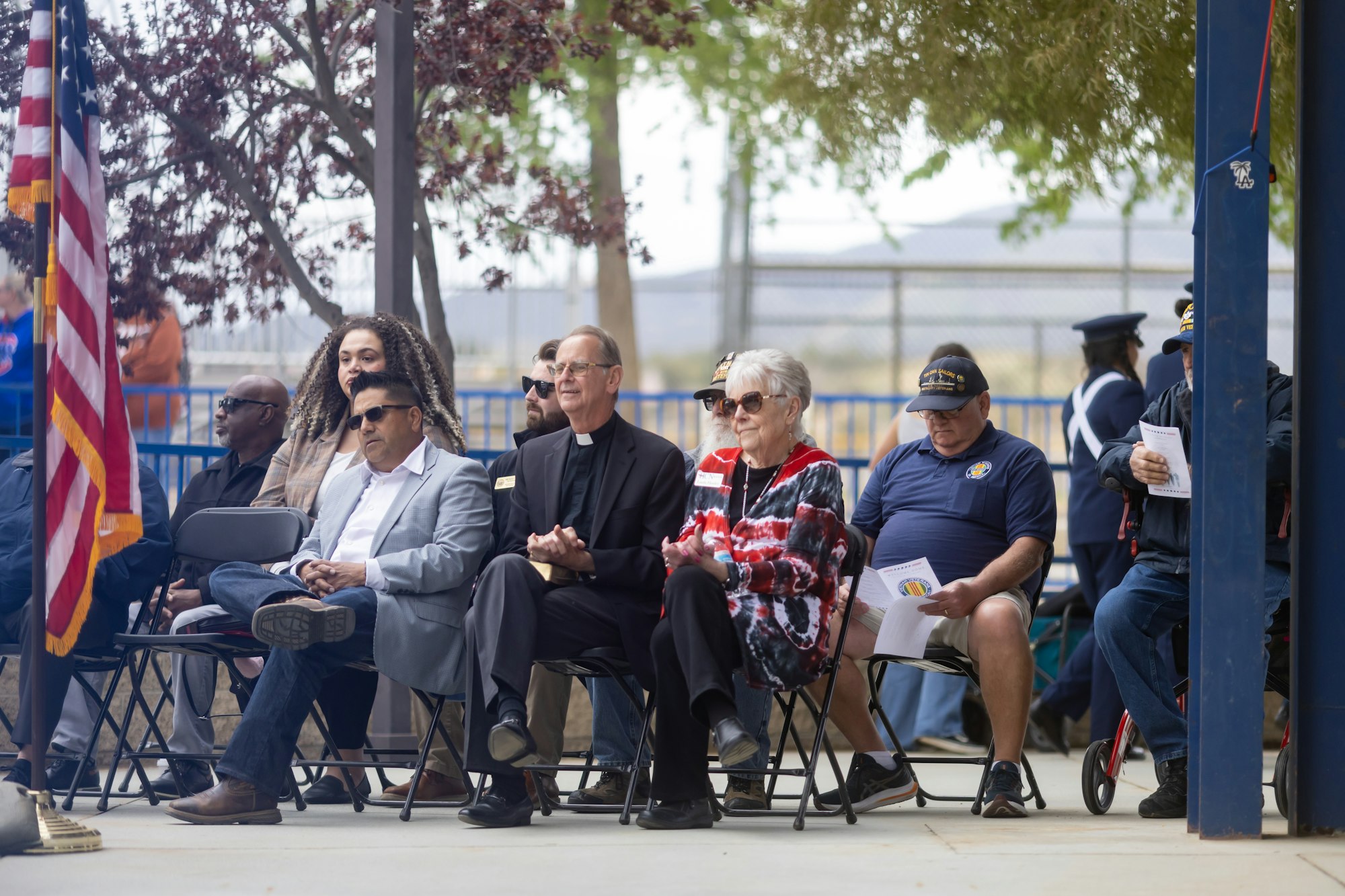 People seated outdoors, some in formal attire. American flag in foreground, some holding papers. Trees and fence in background.