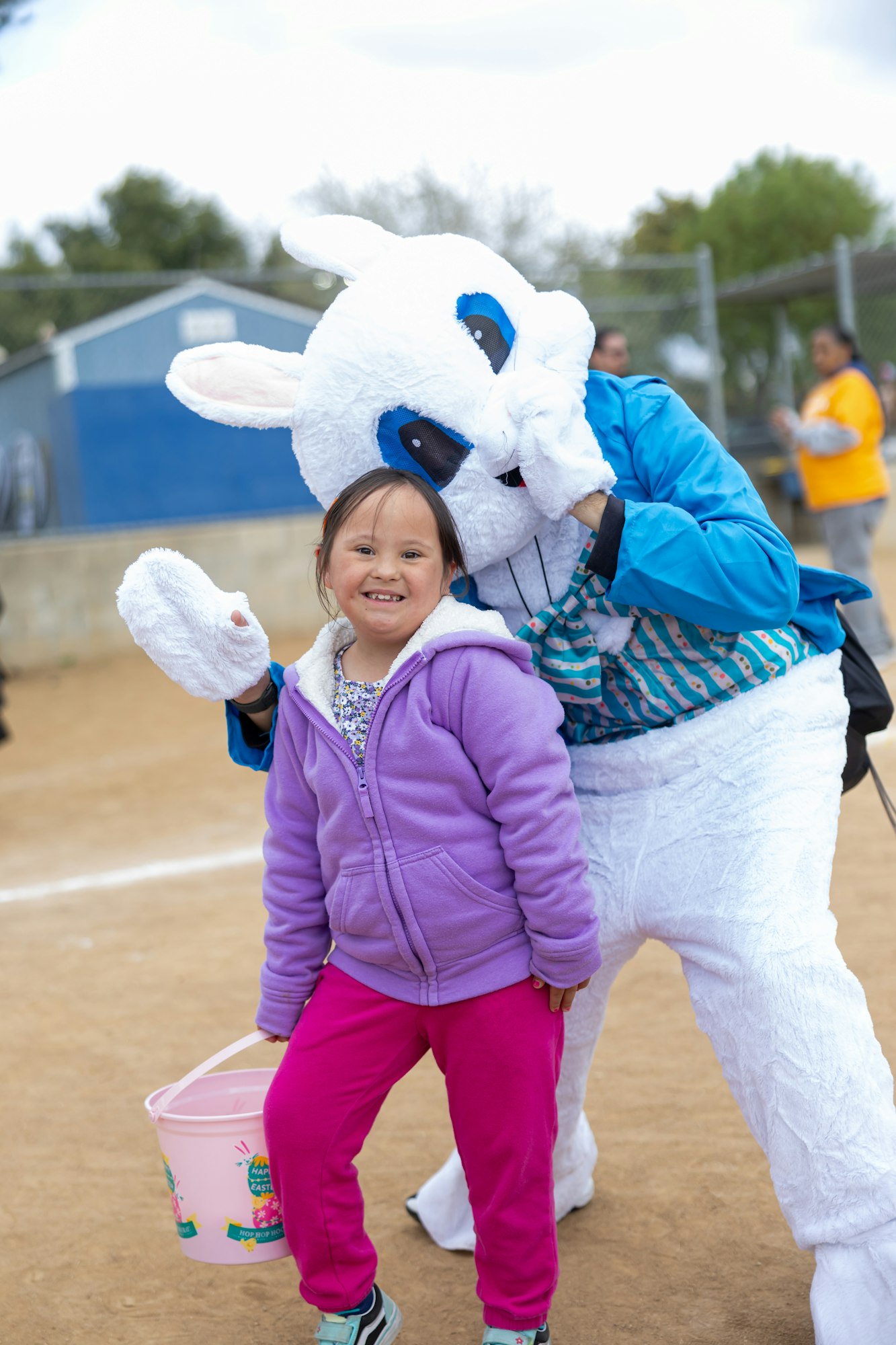 A child in colorful clothing smiles while posing with a person in a bunny costume, holding an Easter-themed bucket.