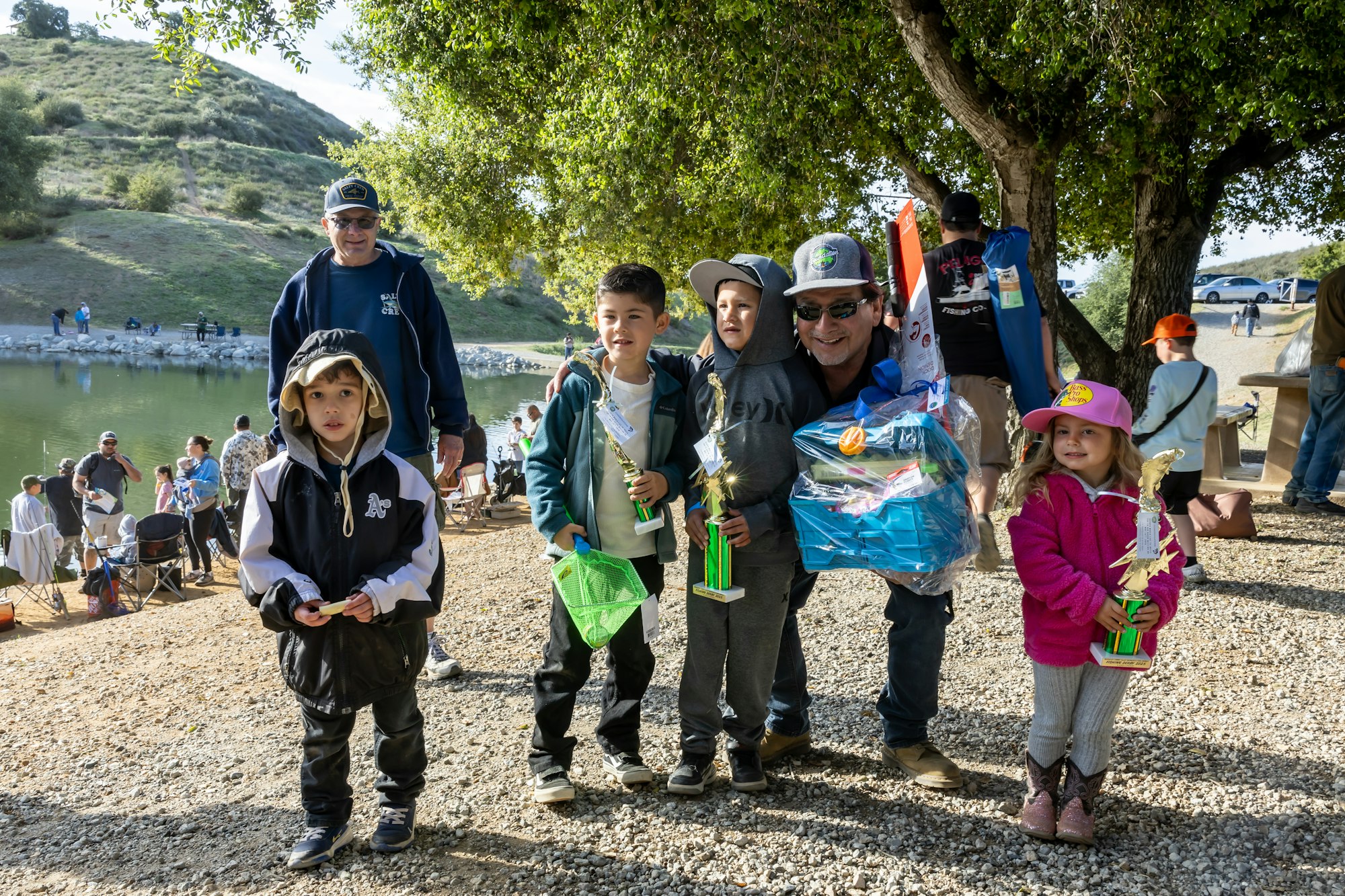 A group of children and adults holding trophies and prizes near a lake, surrounded by trees and grassy hills.