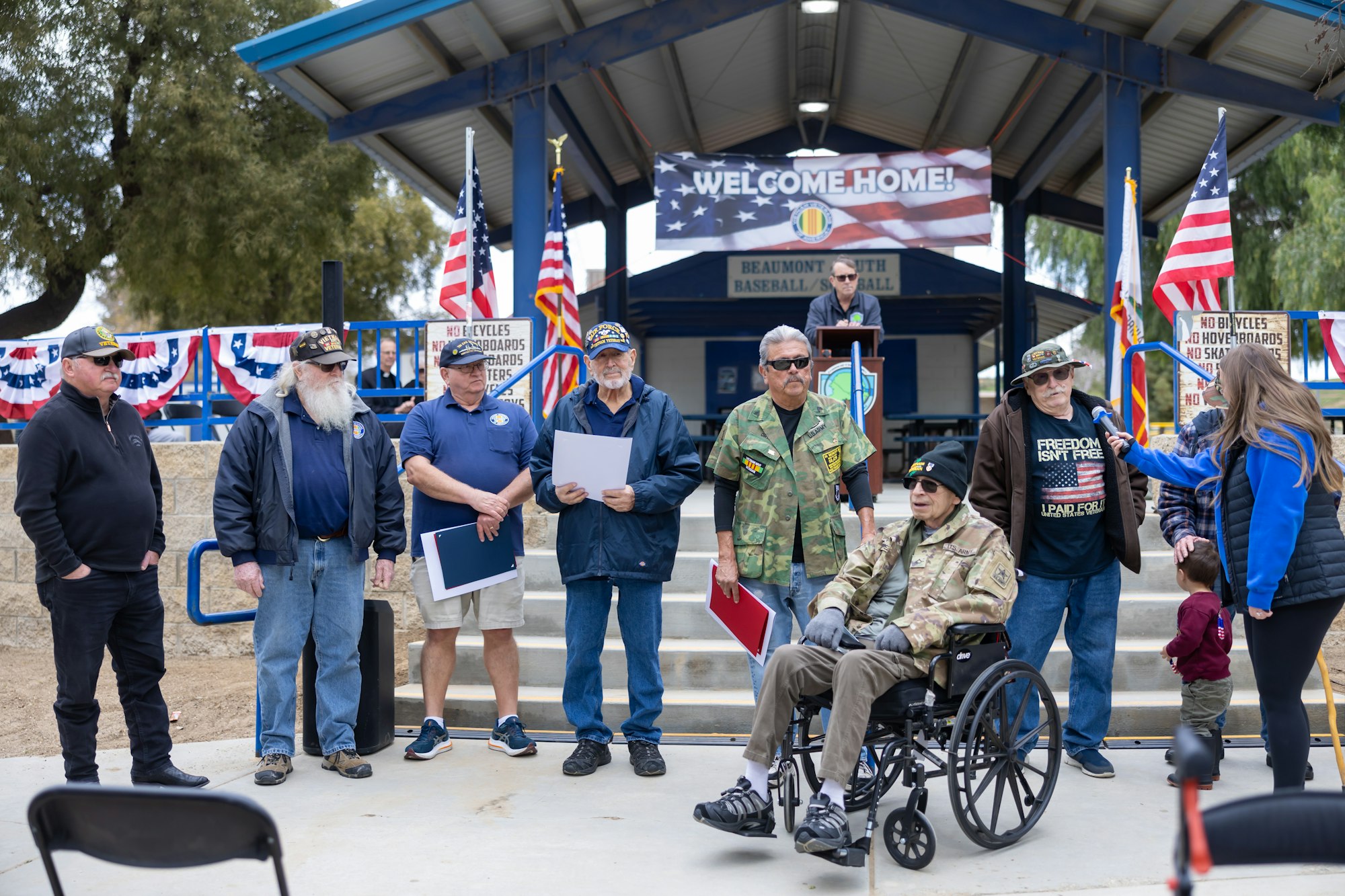 A group of veterans at an outdoor welcome event, with American flags, and a person speaking into a microphone.