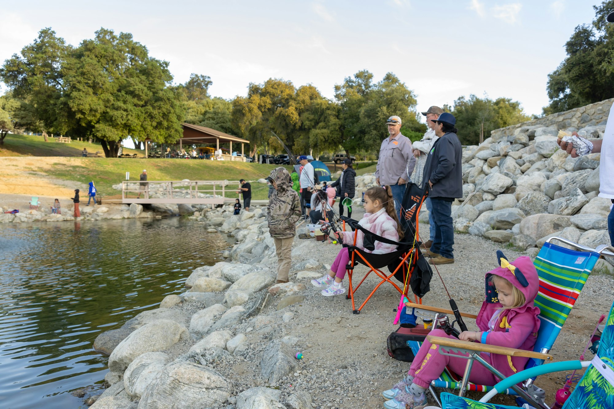 People fishing by a lakeside, with children sitting in chairs and others standing nearby.