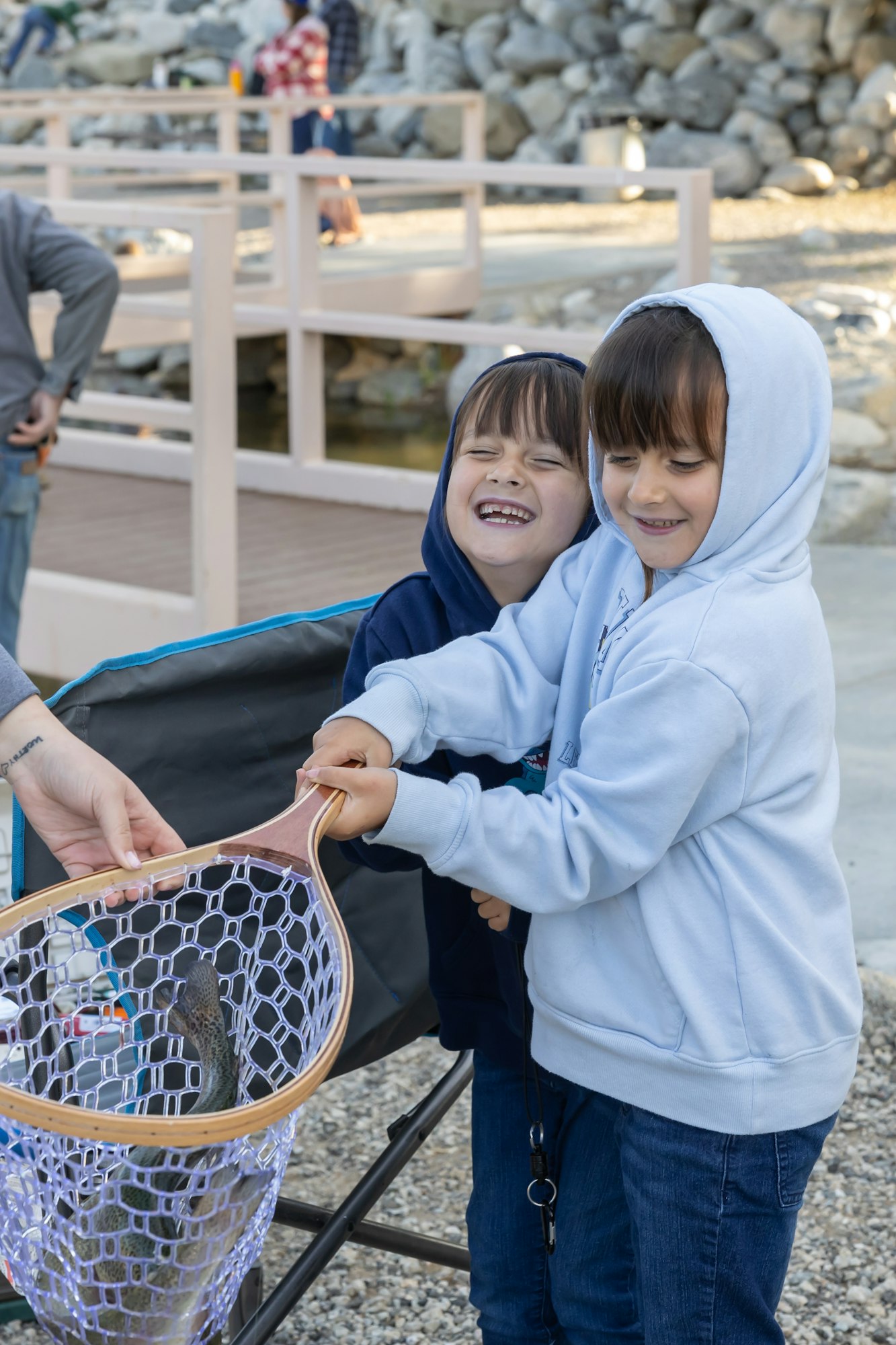Two children in hoodies happily hold a fishing net with a fish inside, while a person assists.