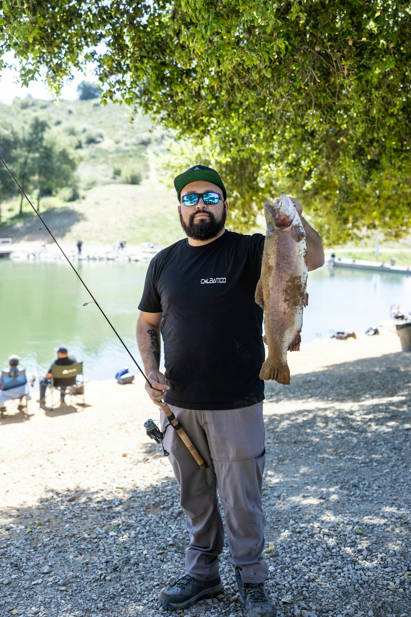 A person in sunglasses holding a large fish and fishing rod near a lakeside, with people relaxing in the background.