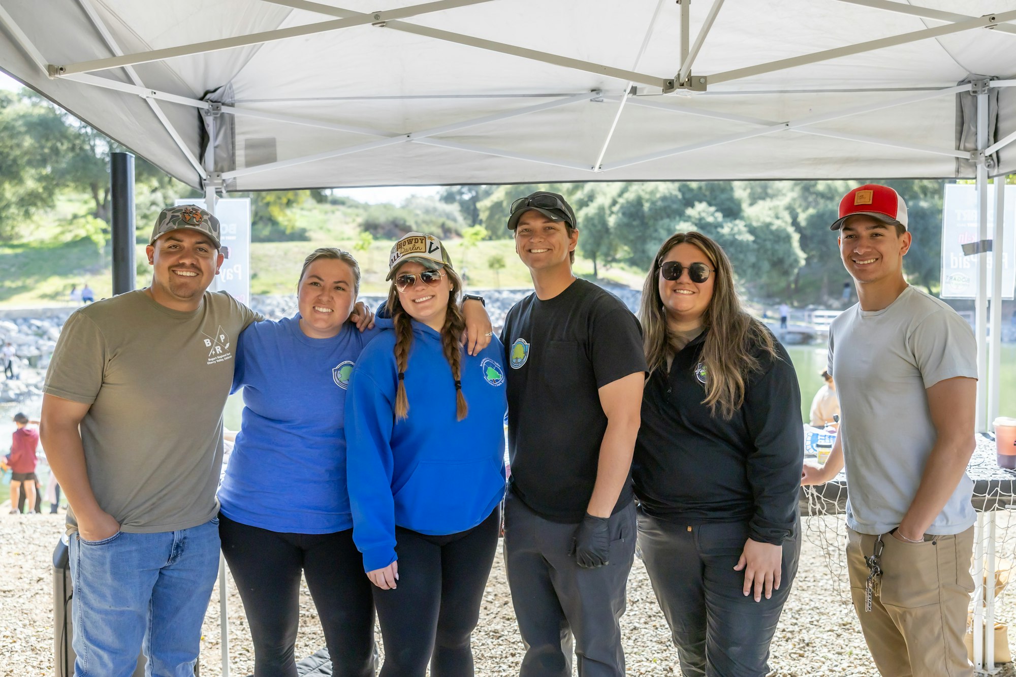 A group of six people smiling under a canopy, wearing casual clothes and hats, outdoors with greenery in the background.