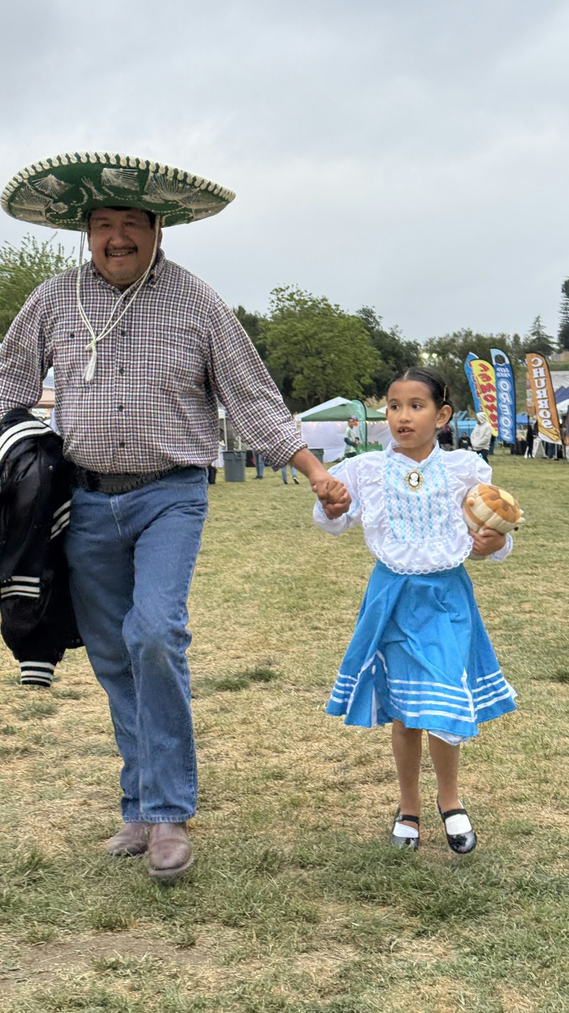 A man and a girl holding hands, outdoors. The man wears a sombrero and the girl is in a traditional dress, holding bread.