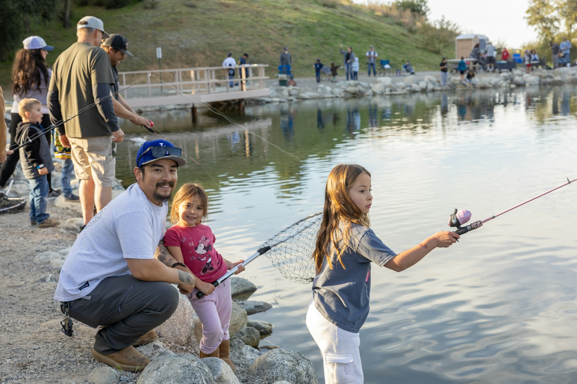A family is fishing by a lake with children holding fishing rods while a group of people gathers in the background.