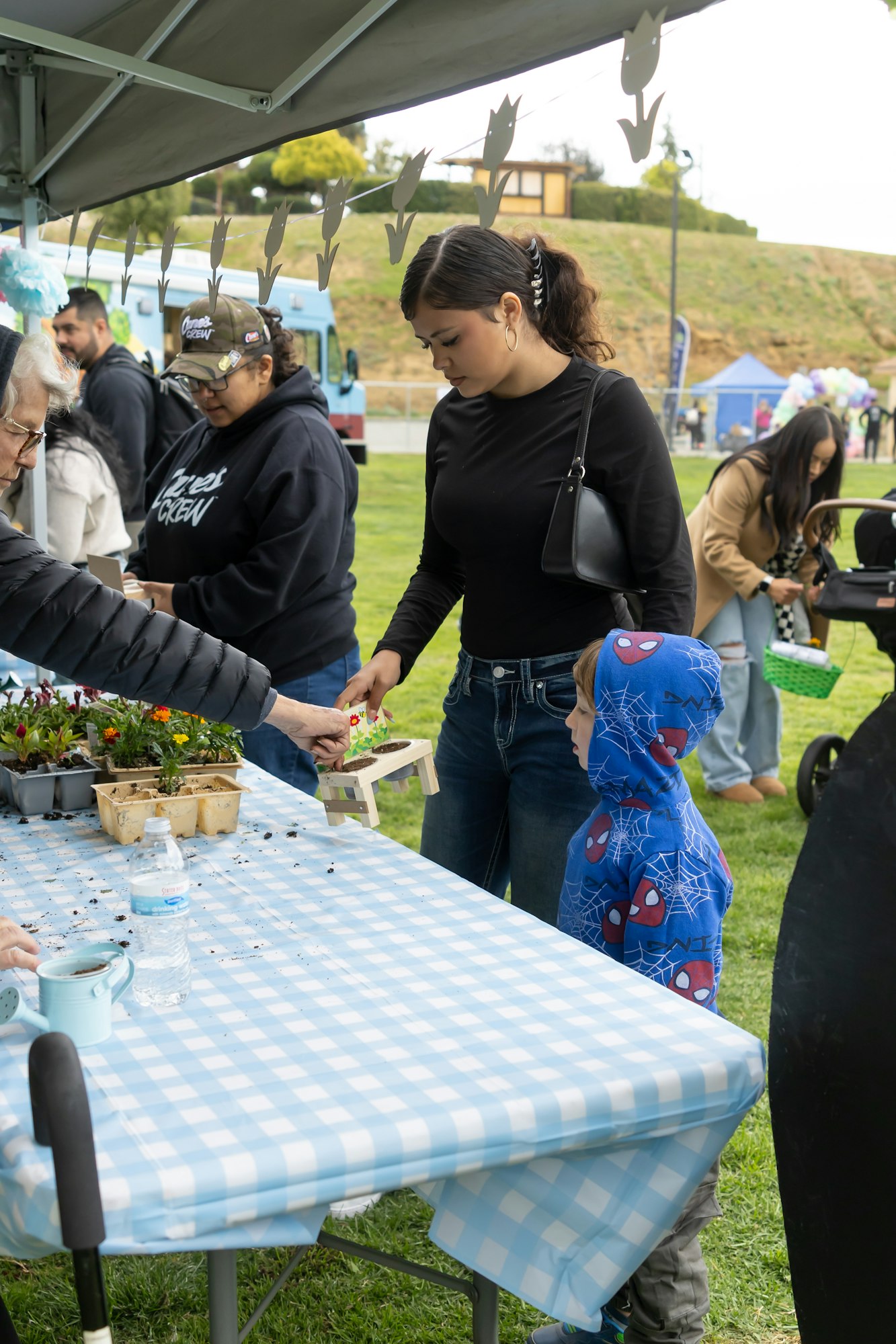 People at a table with plants, a woman hands something to a child in a Spider-Man hoodie at an outdoor market.