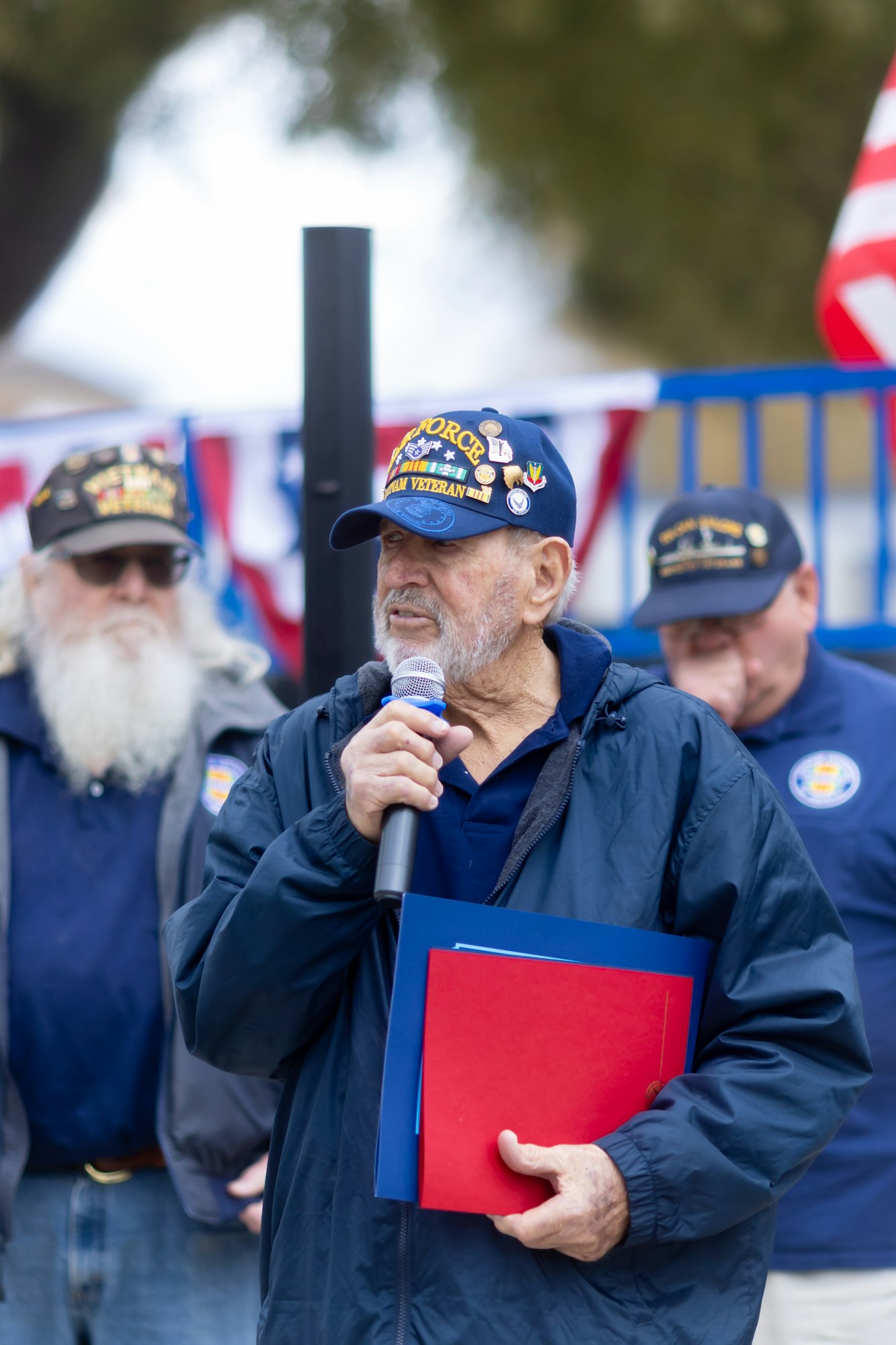 A man in a veteran cap speaking into a microphone, in front of a patriotic backdrop, with others in similar caps nearby.