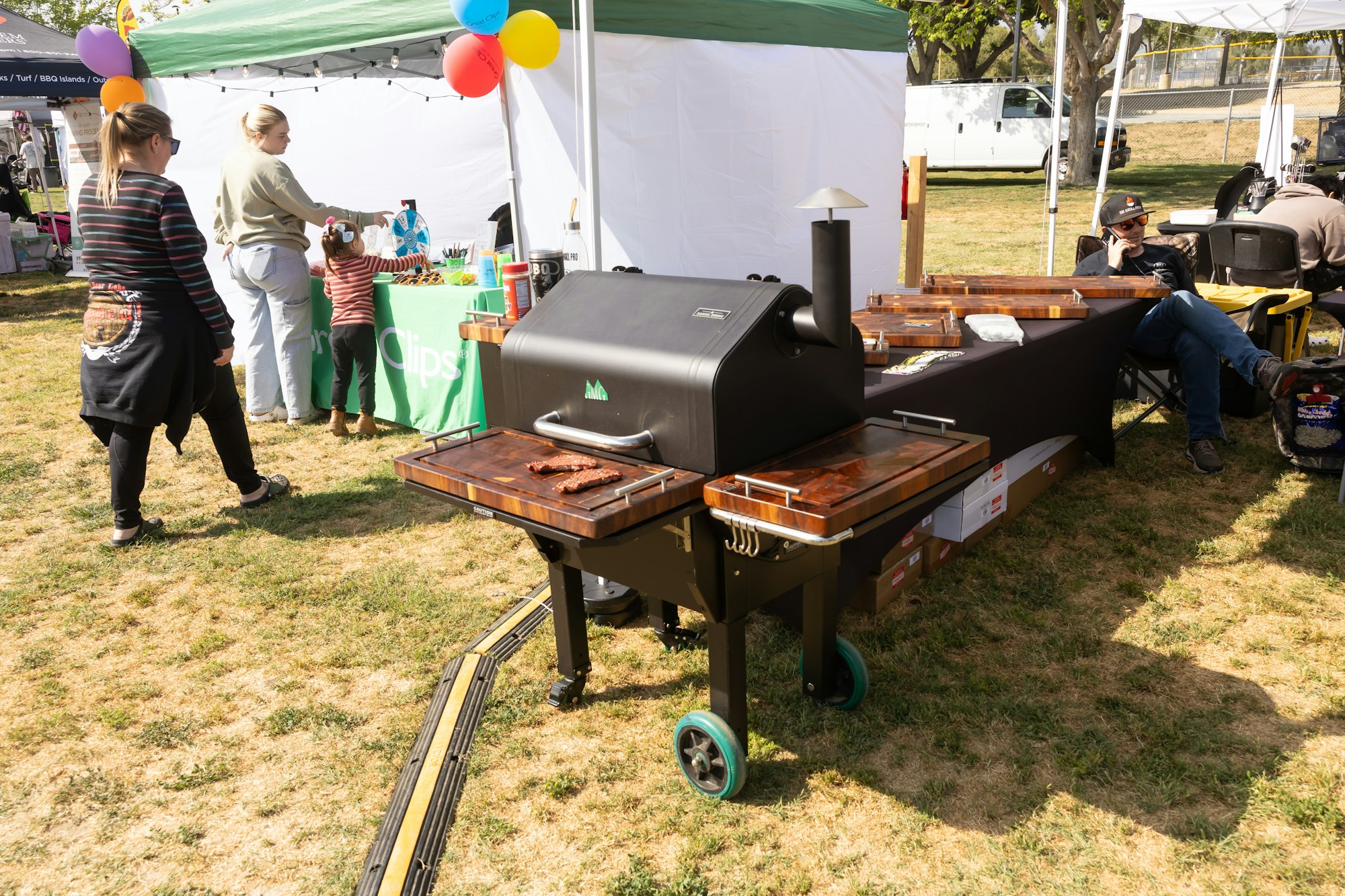 Outdoor fair with a grill, booth, and people interacting; balloons decorate the area.